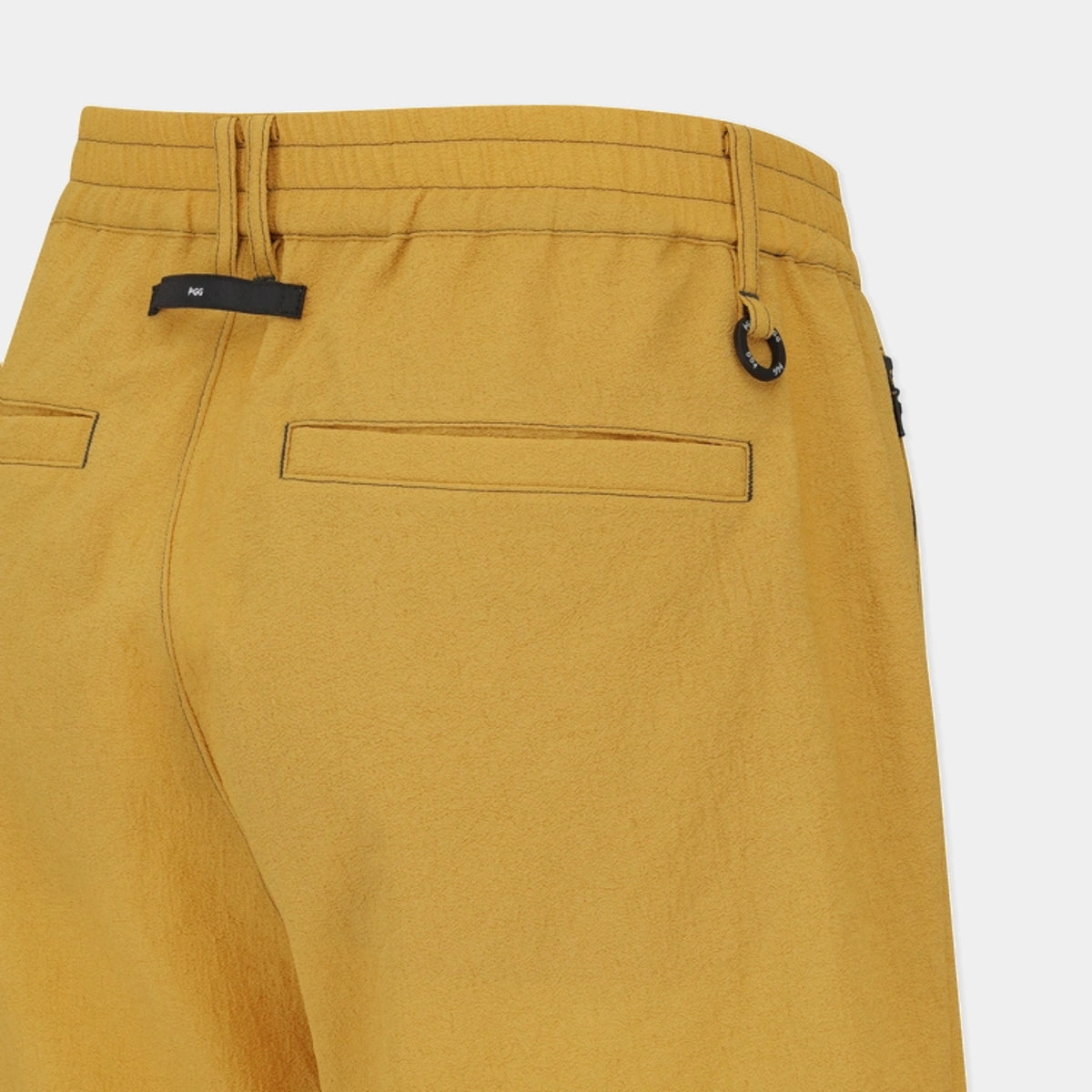 Close-up of the back waistband and rear pockets of mustard yellow trousers, showing a black fabric brand tab and a small black metal ring detail on a light gray background.