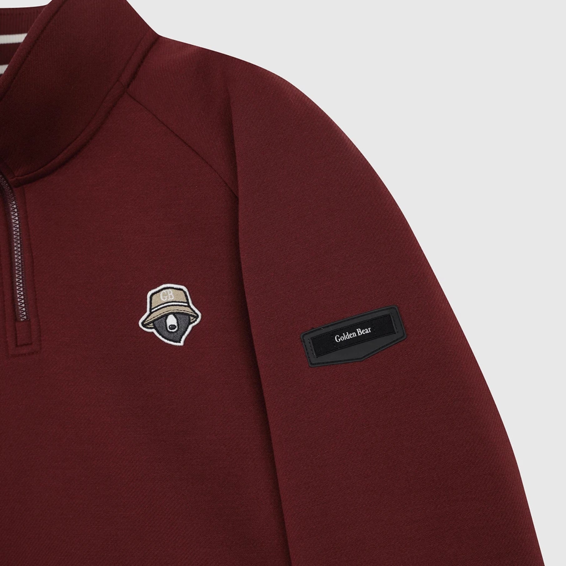 Close-up of a burgundy quarter-zip pullover sweatshirt featuring a black sleeve patch with 'Golden Bear' text and a small embroidered hat logo on the chest, shown against a light gray background.
