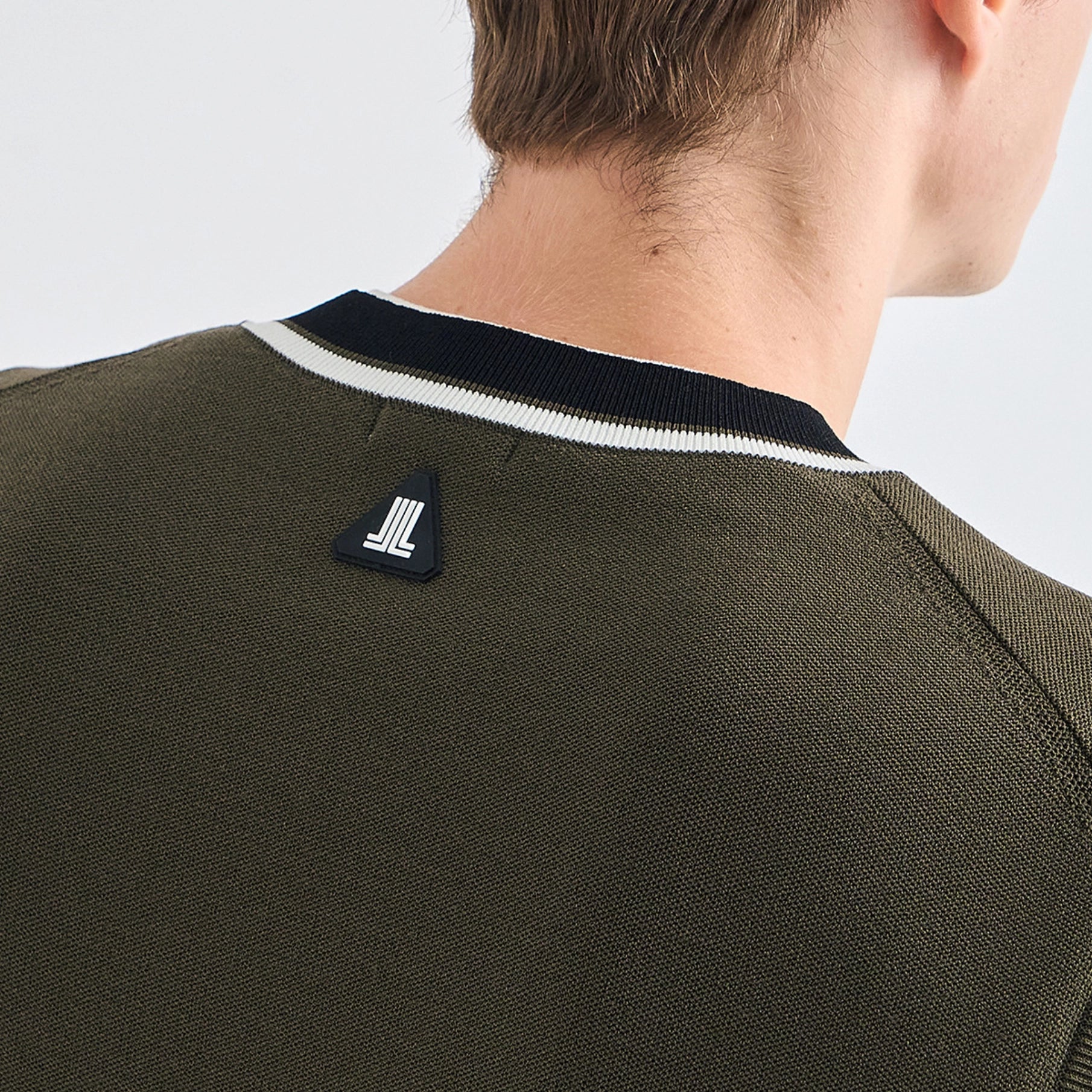 Close-up back view of a person wearing an olive green knit sweater with a black and white striped crew neck and a small black brand patch on the upper back.