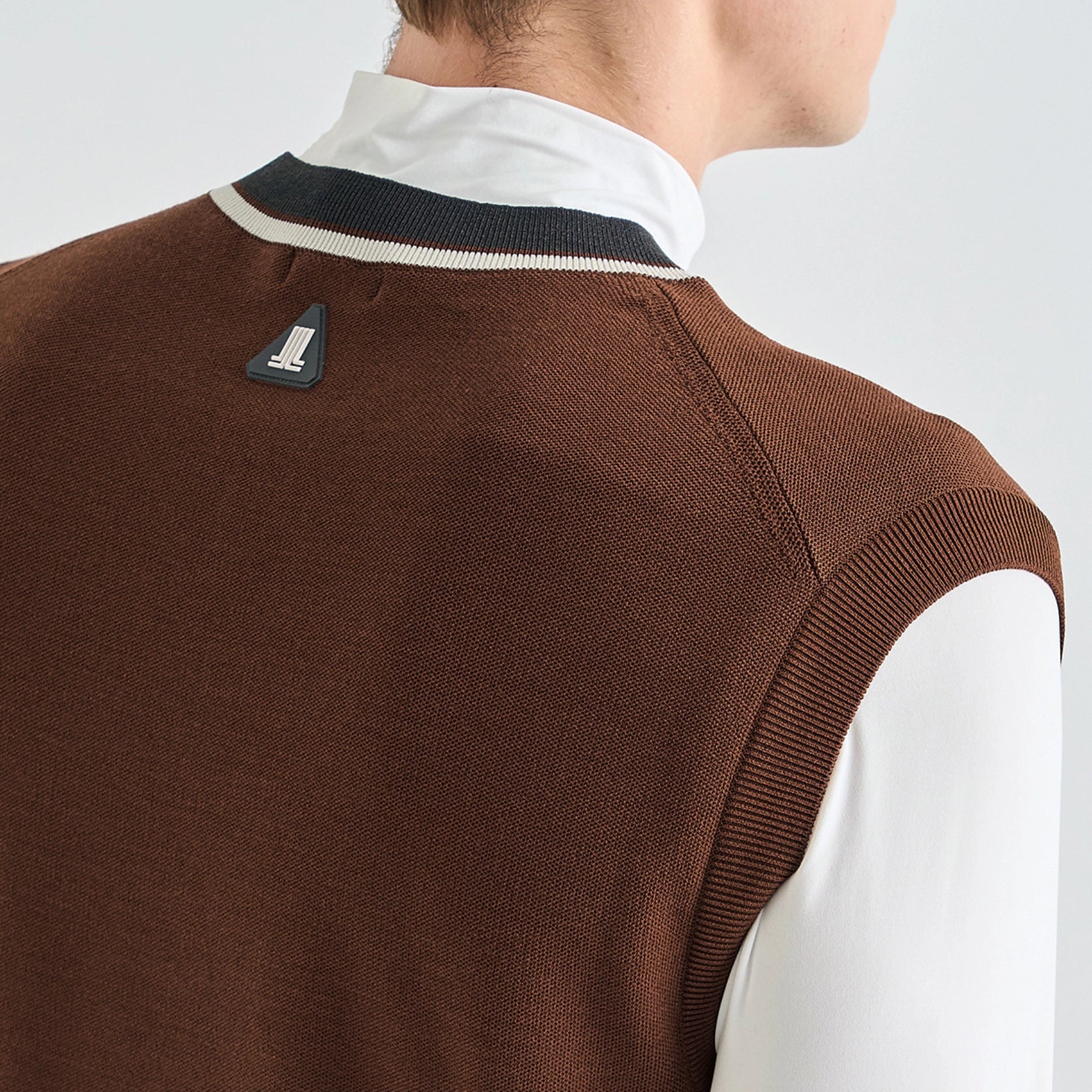 Close-up back view of a person wearing a brown knit sweater vest over a white collared shirt, with a small Sailing Line logo patch near the collar.