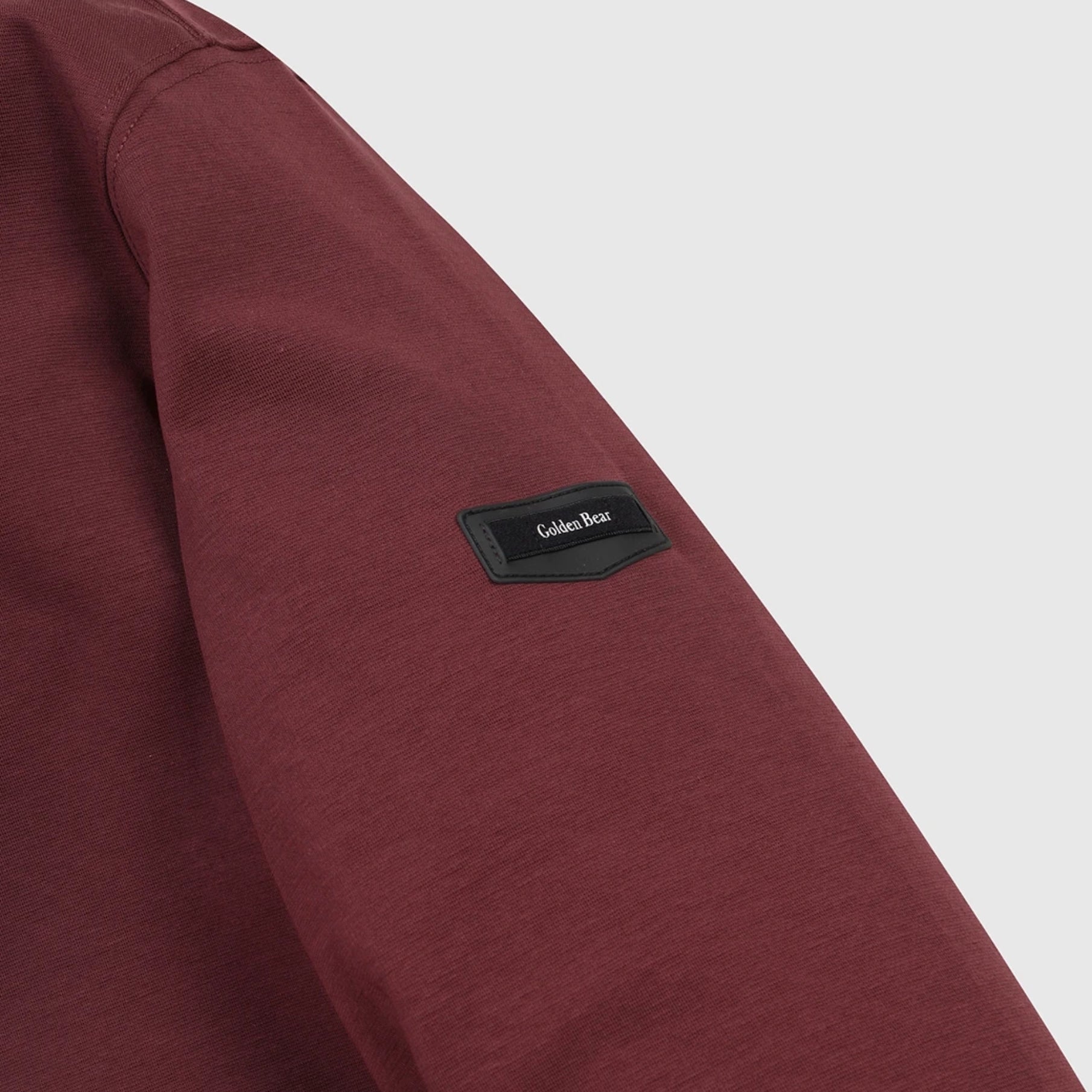 Close-up of a heathered burgundy long-sleeve top on a light gray background, featuring a black rectangular patch on the upper sleeve with the brand name 'Golden Bear' in white background.