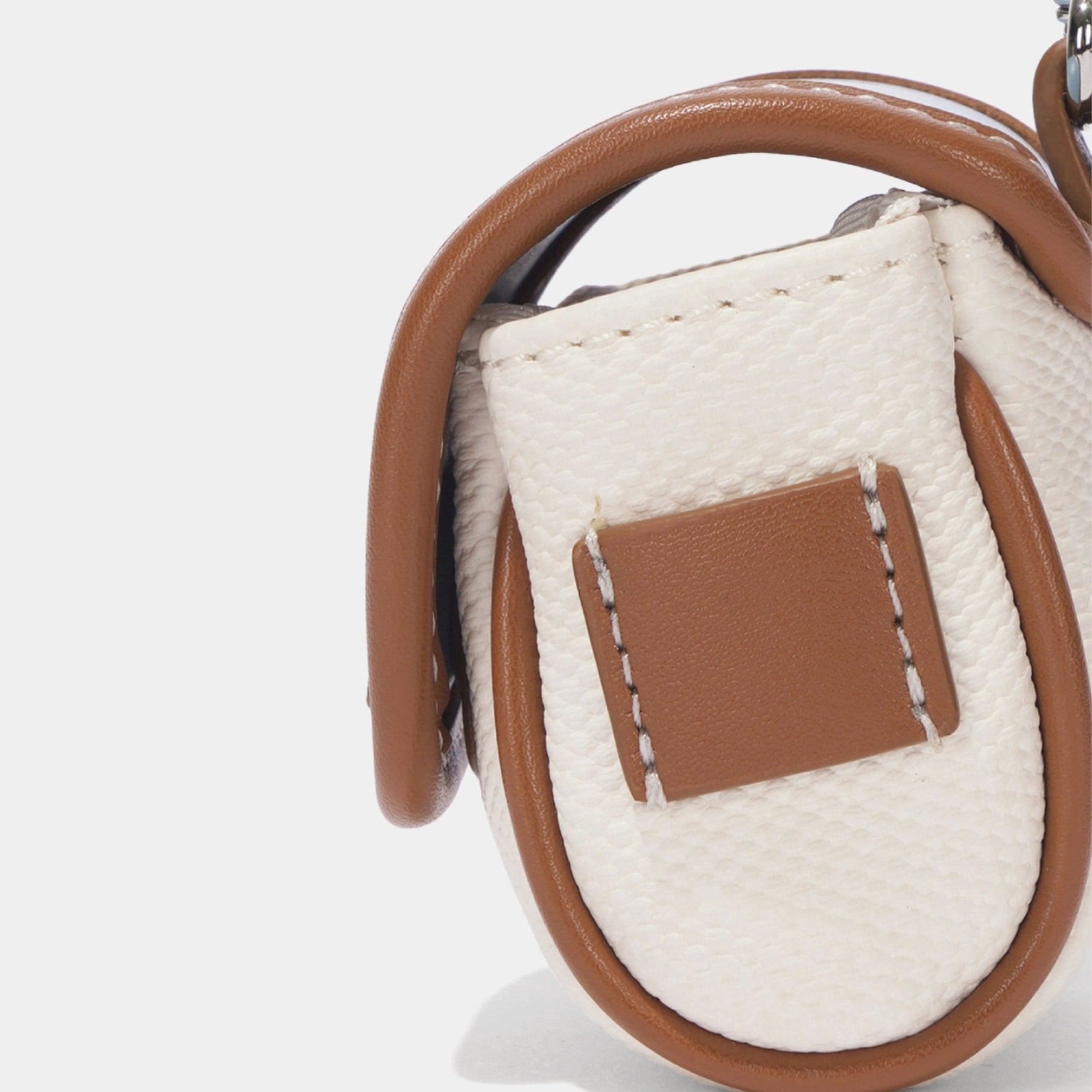Close-up of a handbag in textured off-white woven material with brown leather trim and a matching brown rectangular patch on the front, photographed on a clean white background.