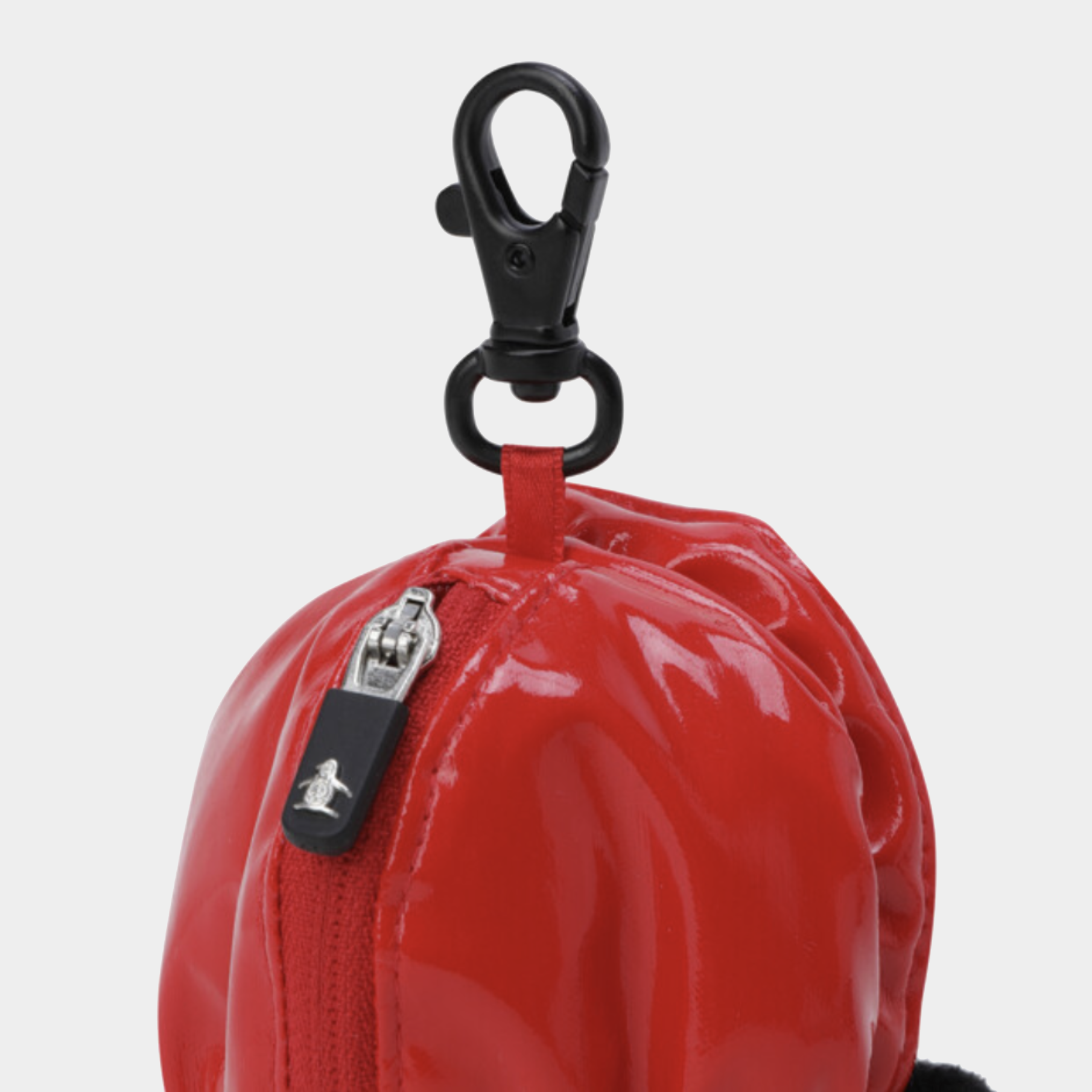 Close-up of a glossy red zippered pouch with a black carabiner clip and a small black tag featuring a white penguin logo, on a light gray background.