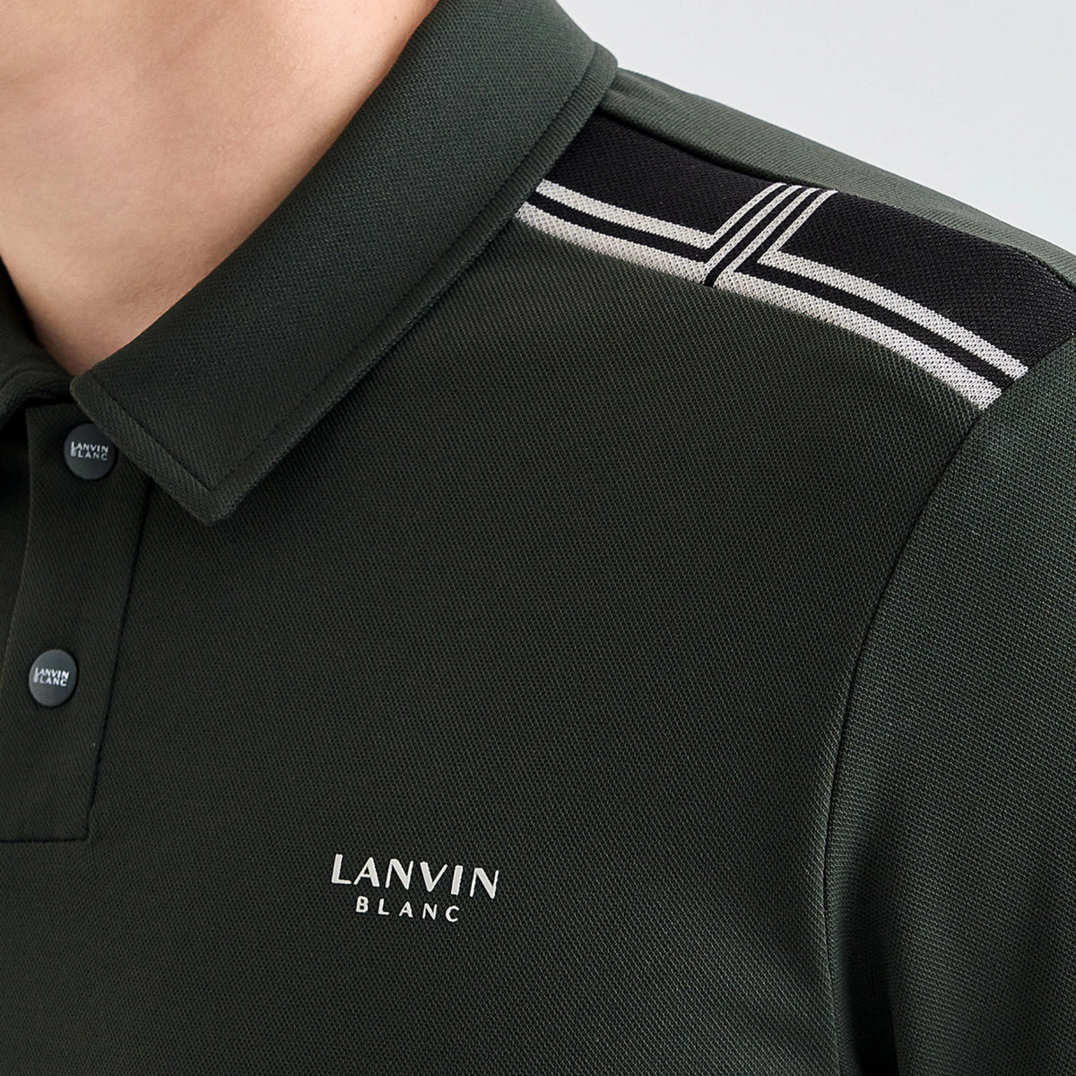 Close-up of a dark green piqué polo shirt worn by a model, featuring a black and white striped shoulder yoke and the white LANVIN BLANC logo on the chest.