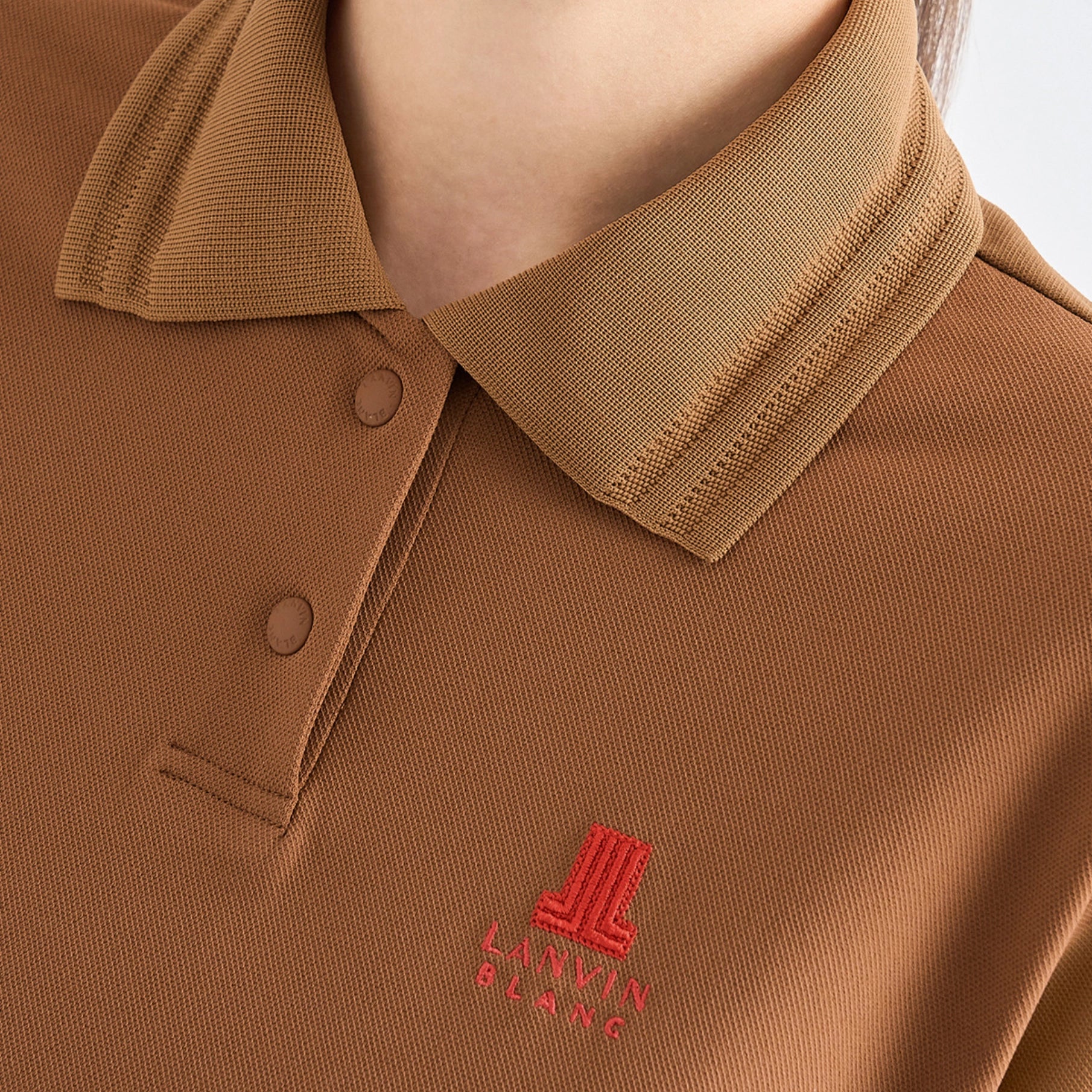 Close-up of a person wearing a brown polo shirt with a matching collar and a small red embroidered logo on the chest.