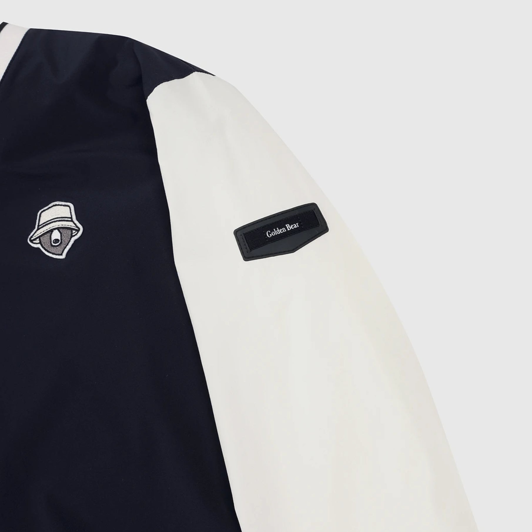 Close-up of a color-blocked jacket sleeve with a dark navy body, an off-white sleeve, a black 'Cold-Weather' patch, and a white embroidered bear logo patch.