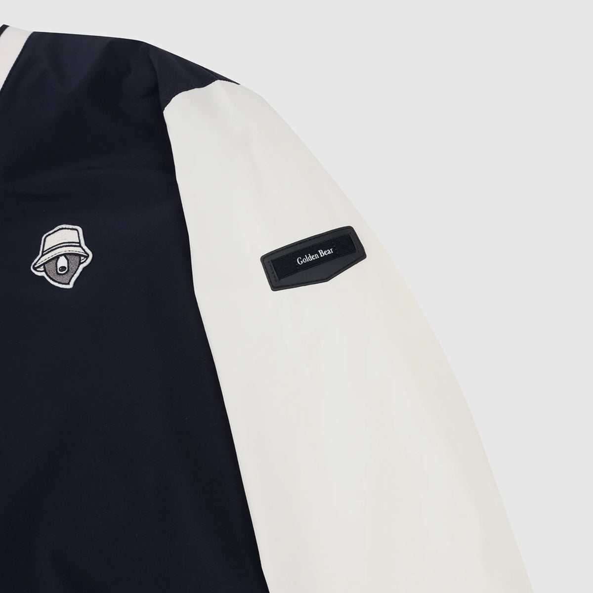 Close-up of a color-blocked jacket sleeve with a dark navy body, an off-white sleeve, a black 'Cold-Weather' patch, and a white embroidered bear logo patch.