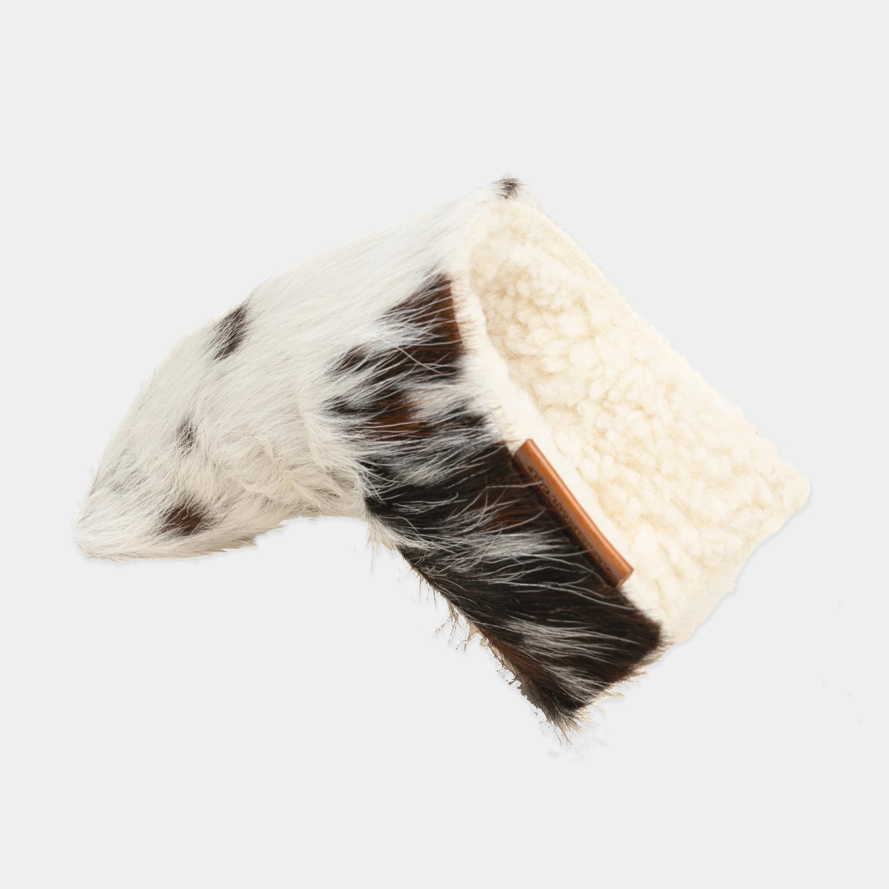 A putter cover featuring one half made of black and white cowhide fur and the other half made of cream-colored sherpa fleece, with a small brown leather tab on the sherpa side.
