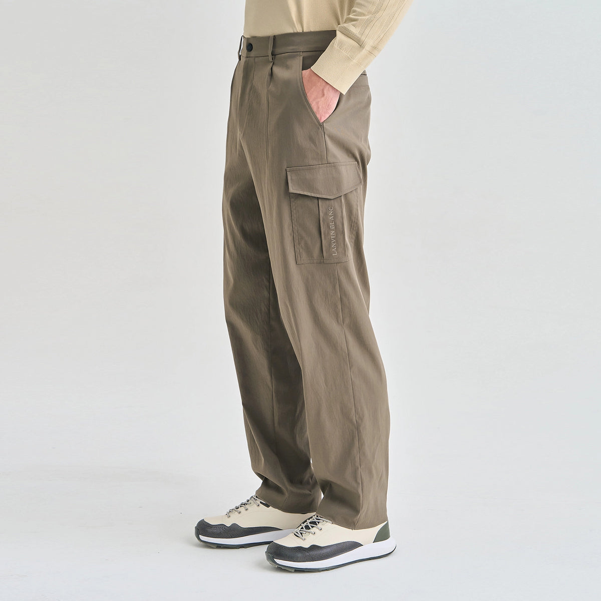 A person wearing loose-fit cargo pants in a taupe-brown color, paired with a beige long-sleeve top and beige-and-black sneakers, shown from the waist down against a light gray studio background.