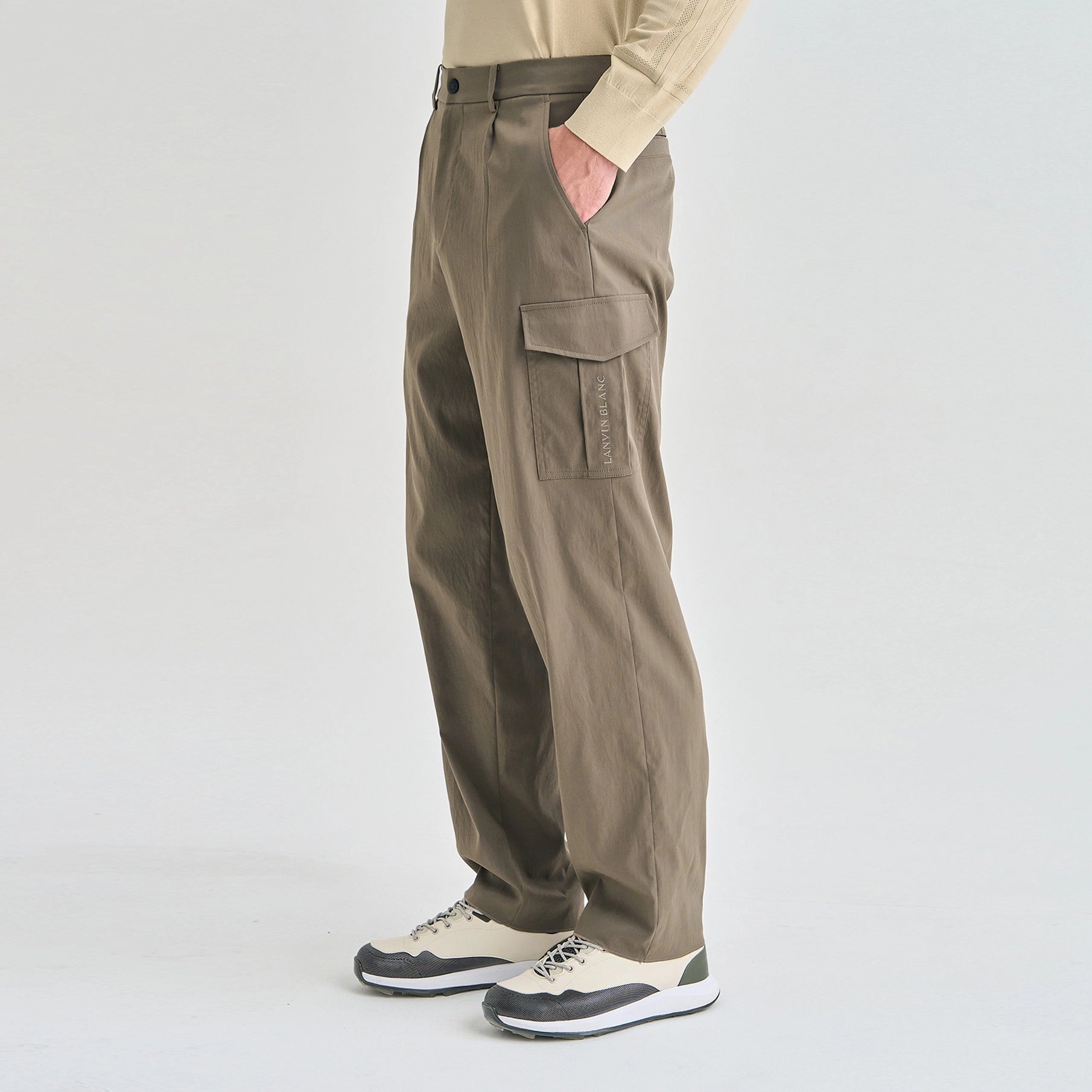 A person wearing loose-fit cargo pants in a taupe-brown color, paired with a beige long-sleeve top and beige-and-black sneakers, shown from the waist down against a light gray studio background.
