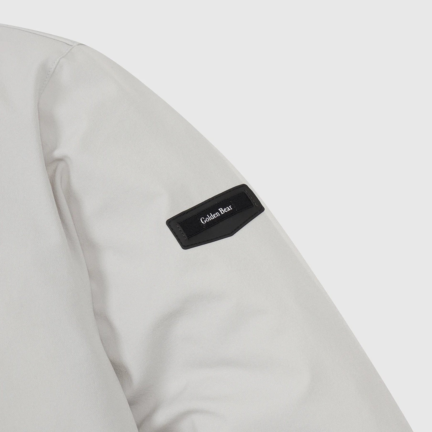 Close-up of a light gray jacket sleeve featuring a black fabric patch with the brand name 'Golden Bear' in white text.