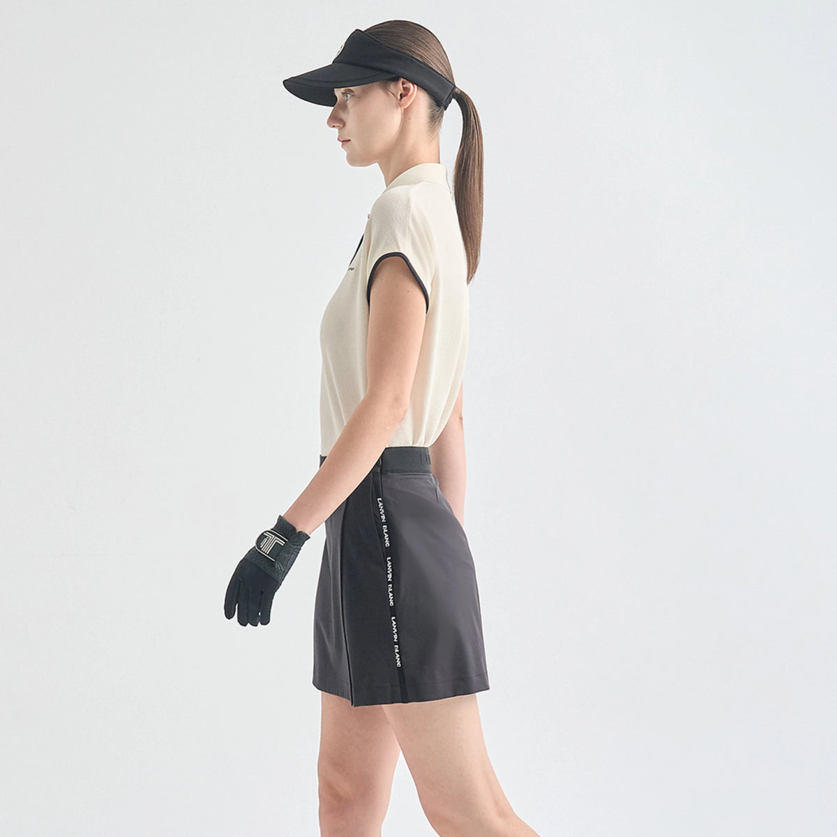A woman in side profile wearing a black visor, cream sleeveless athletic top, dark gray athletic shorts with white text detailing, and a black glove, set against a plain light gray studio background.