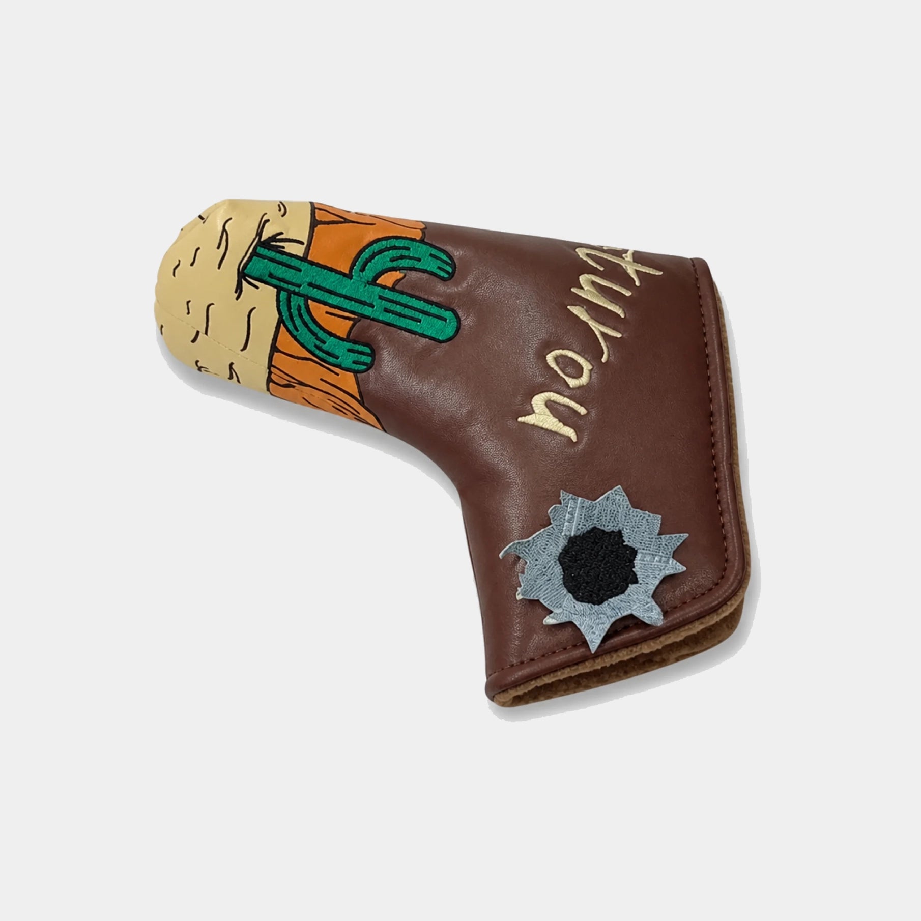 A brown leather golf putter head cover featuring a desert illustration with a green cactus, orange rock, and a silver bullet-shaped patch with a black center, on a light gray background.