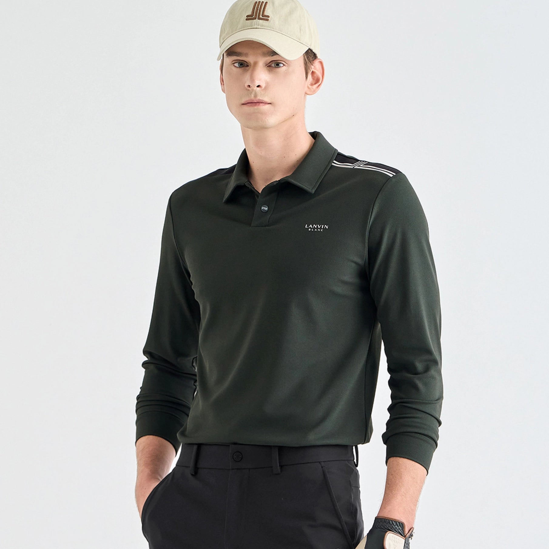 A man wearing a dark forest green long-sleeve polo shirt with white shoulder stripes and a small 'LANVIN BLANC' logo on the chest, paired with a beige cap and black trousers, against a light gray studio background.
