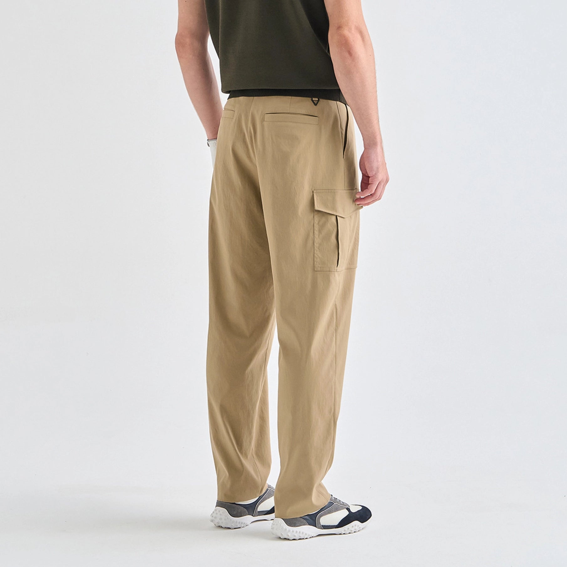 A person shown from the back wearing loose tan cargo pants, a dark olive green shirt, and gray and white sneakers against a clean light gray studio background.