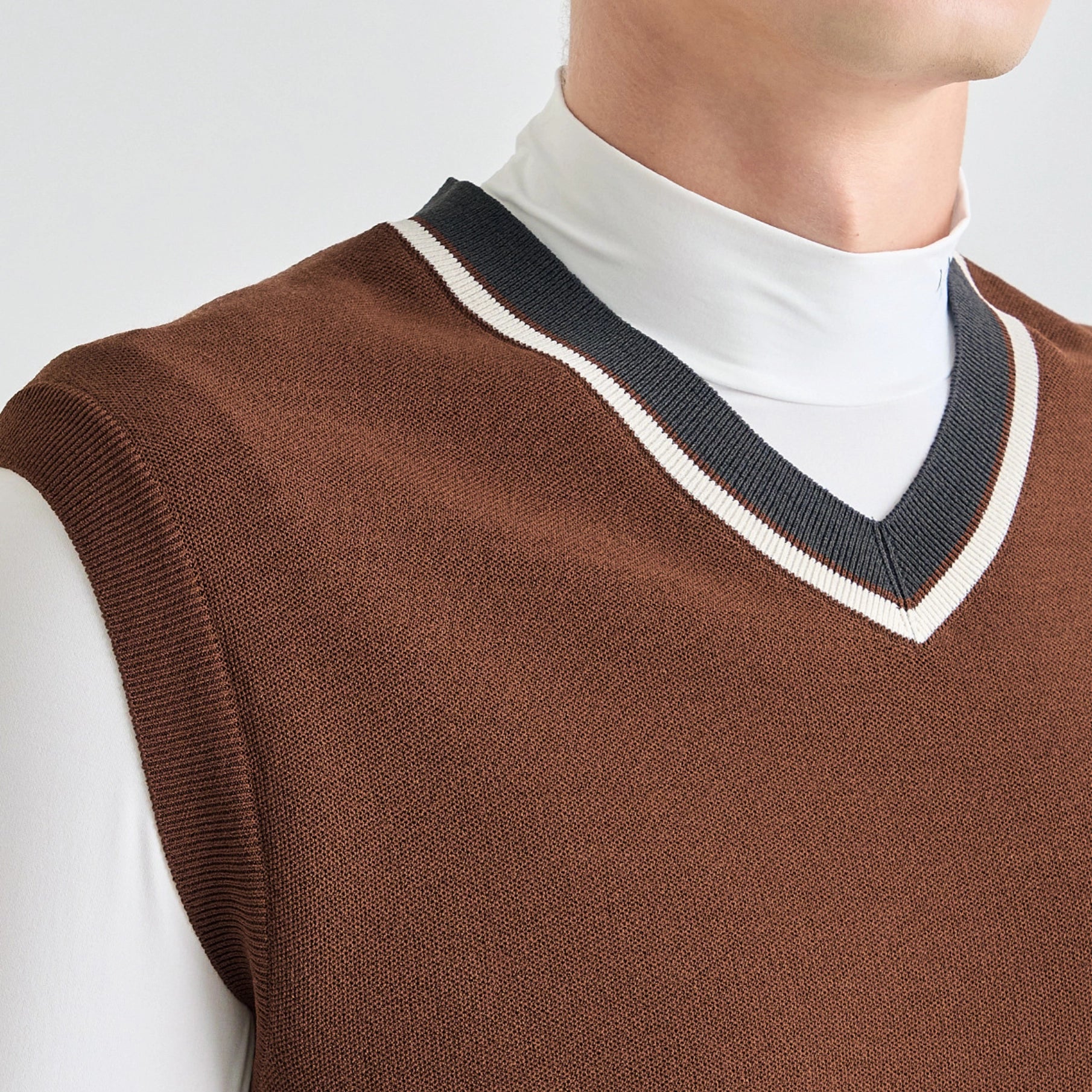 A close-up of a brown knit sweater vest worn over a white mock neck shirt. The vest features a V-neck collar with a dark navy blue ribbed trim and a thin cream stripe.