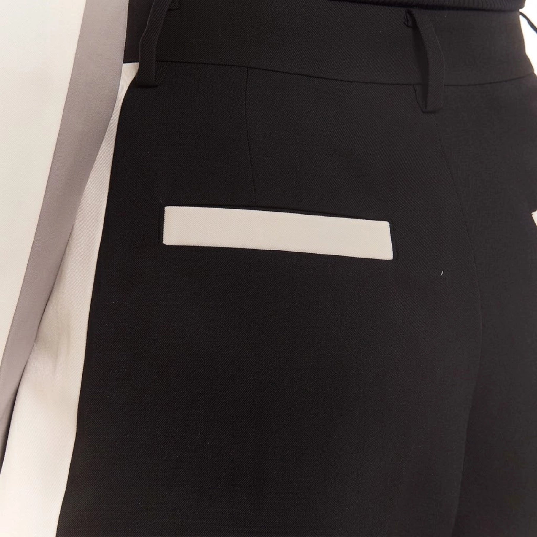 Close-up of black tailored trousers on a mannequin, showing the waistband, belt loops, and a contrasting white rectangular patch pocket.