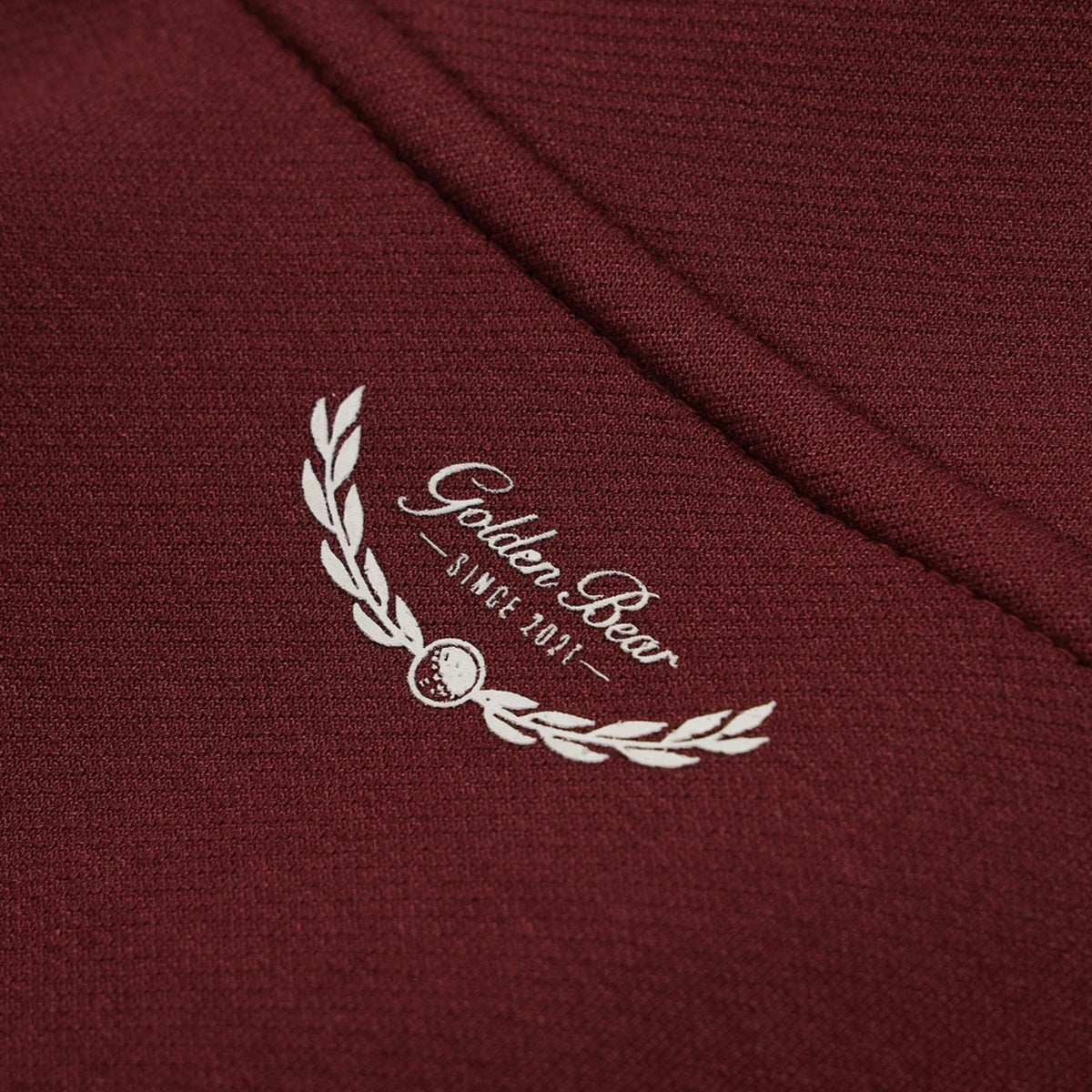 Close-up of a burgundy knit fabric with a white embroidered laurel wreath and the words 'Golden Bear' and 'SINCE 2021'.