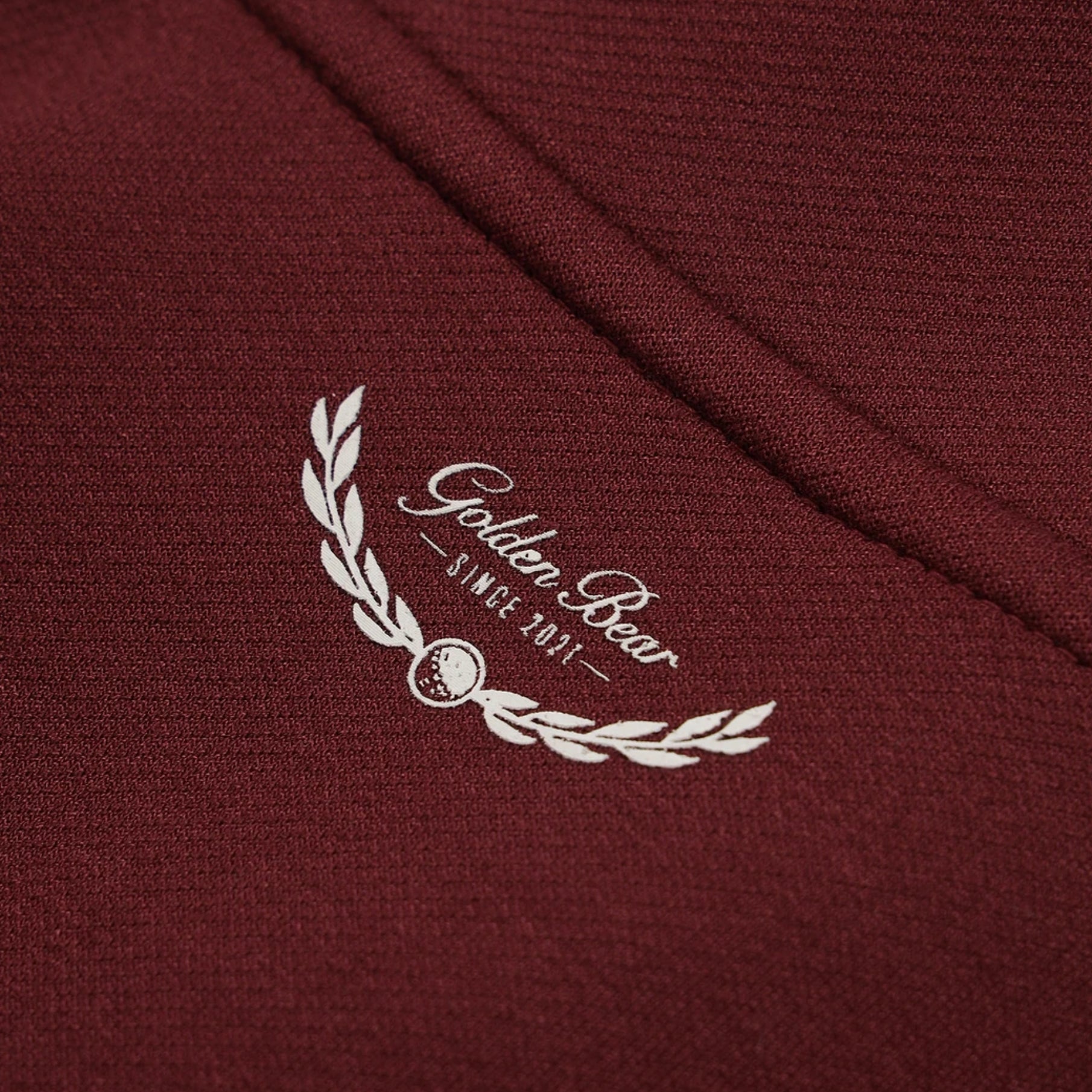 Close-up of a burgundy knit fabric with a white embroidered laurel wreath and the words 'Golden Bear' and 'SINCE 2021'.