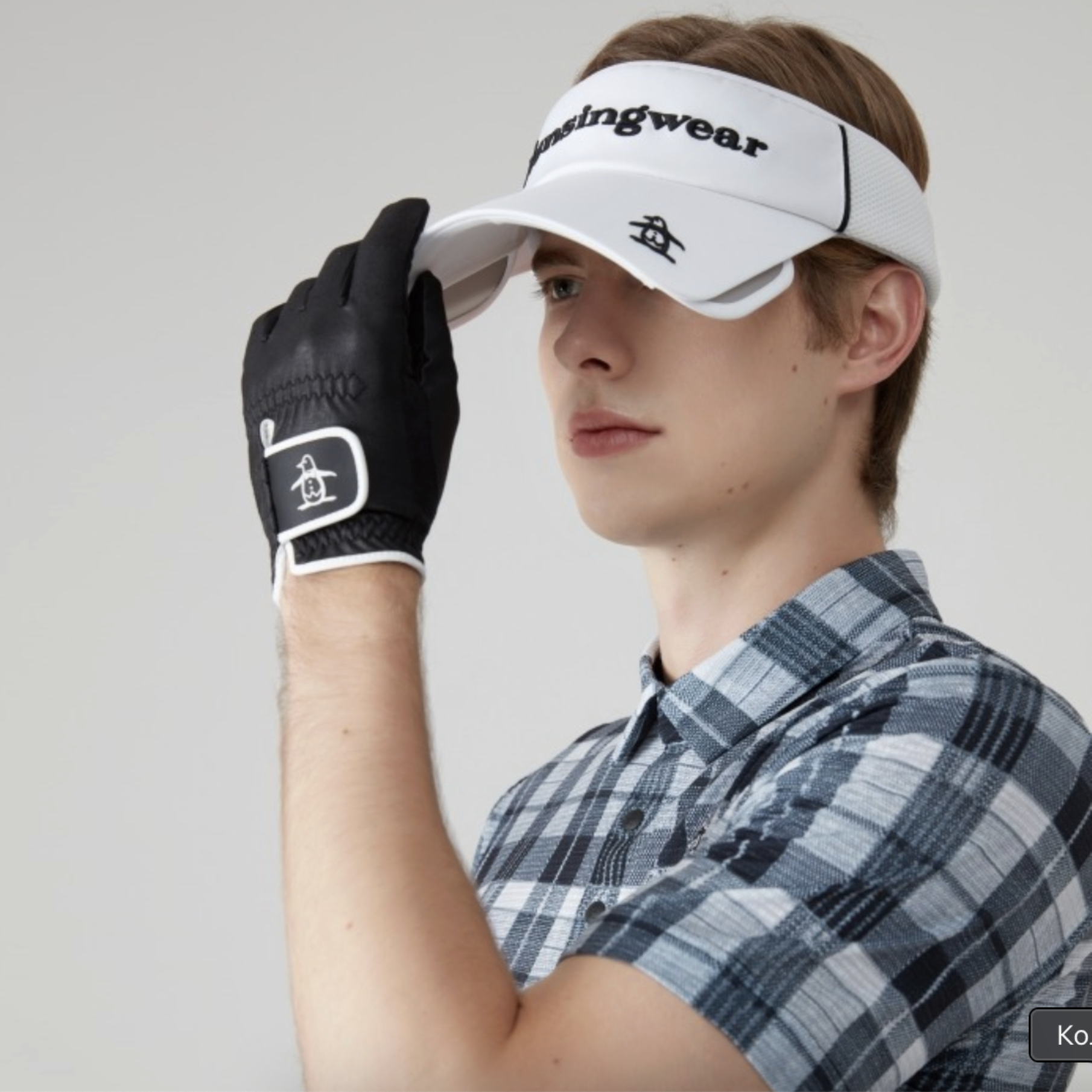 A man in a blue and white plaid shirt wearing a white visor with black text, a black golf glove, and a gray background.