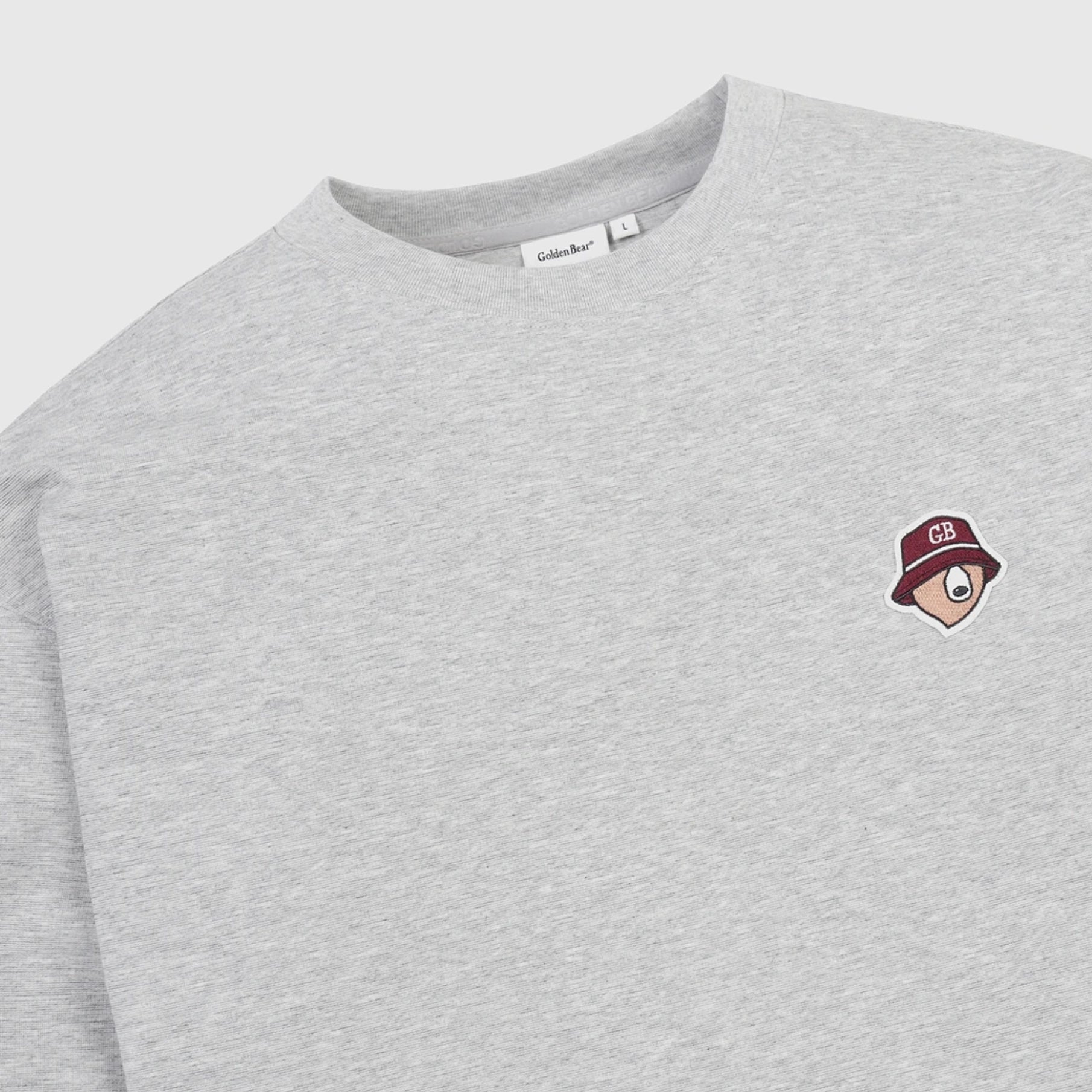 A close-up of a light heather gray crewneck sweatshirt featuring a small embroidered chest graphic of a cartoon face wearing a maroon bucket hat with 'GB' on it. The brand name 'Glockner' is visible on the inner neck label.