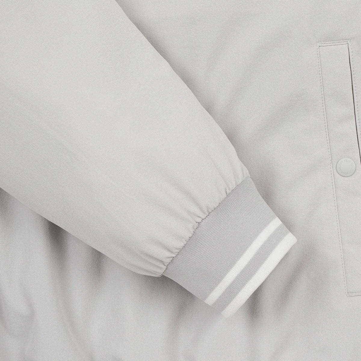 Close-up of a light gray jacket sleeve with a ribbed cuff featuring two white stripes, next to a matching buttoned pocket.
