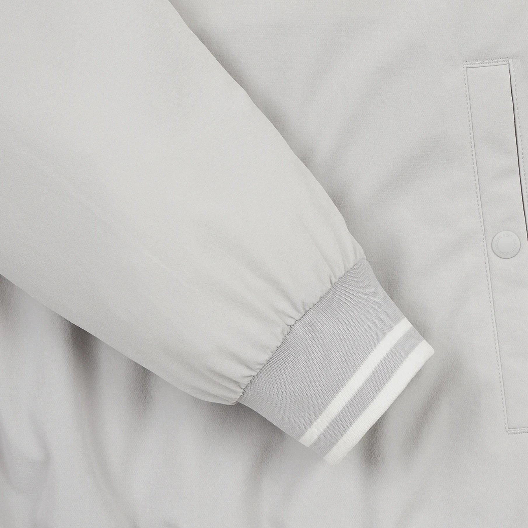 Close-up of a light gray jacket sleeve with a ribbed cuff featuring two white stripes, next to a matching buttoned pocket.