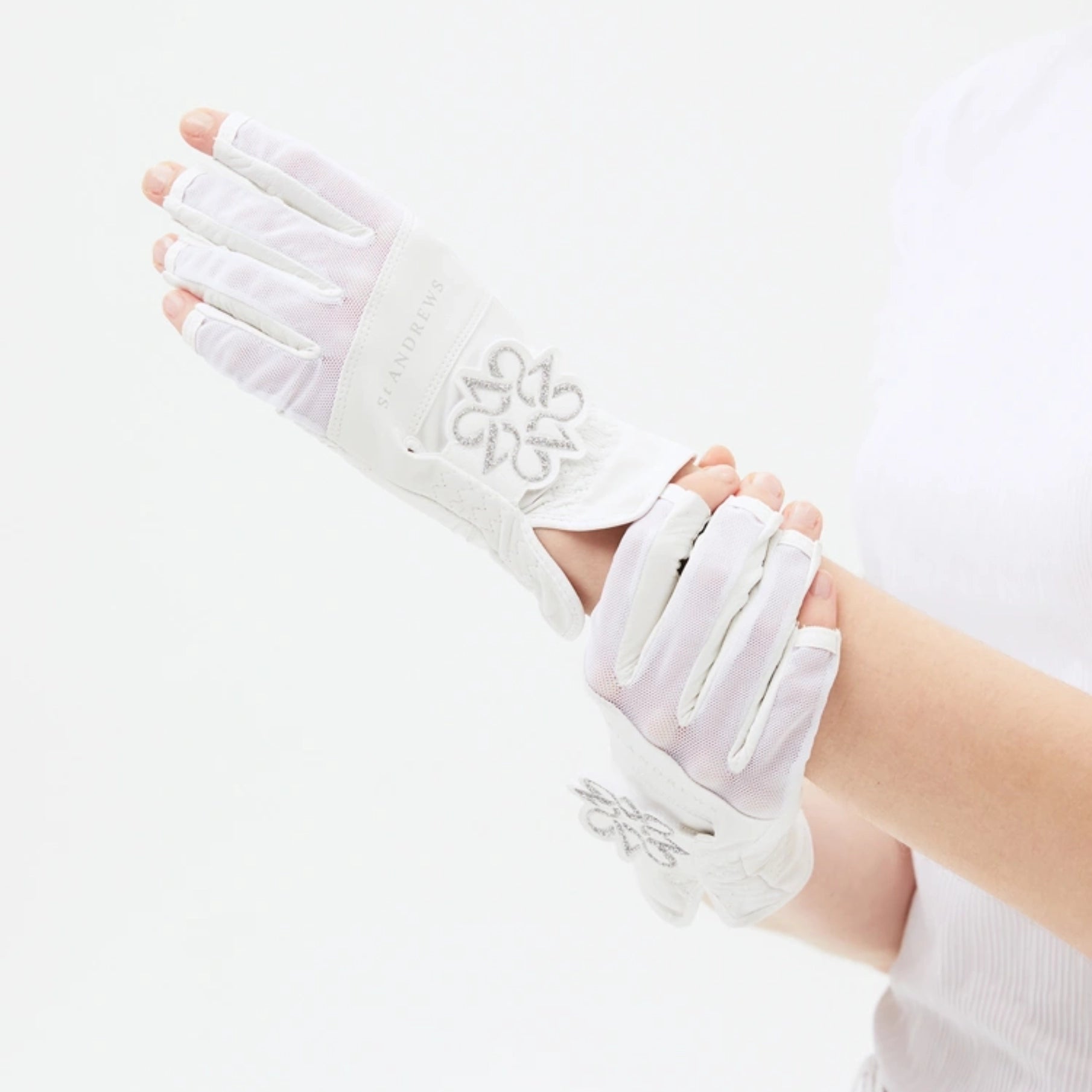 White fingerless gloves with silver floral embroidery and a debossed brand name, shown being worn on a person's hands against a clean white background.