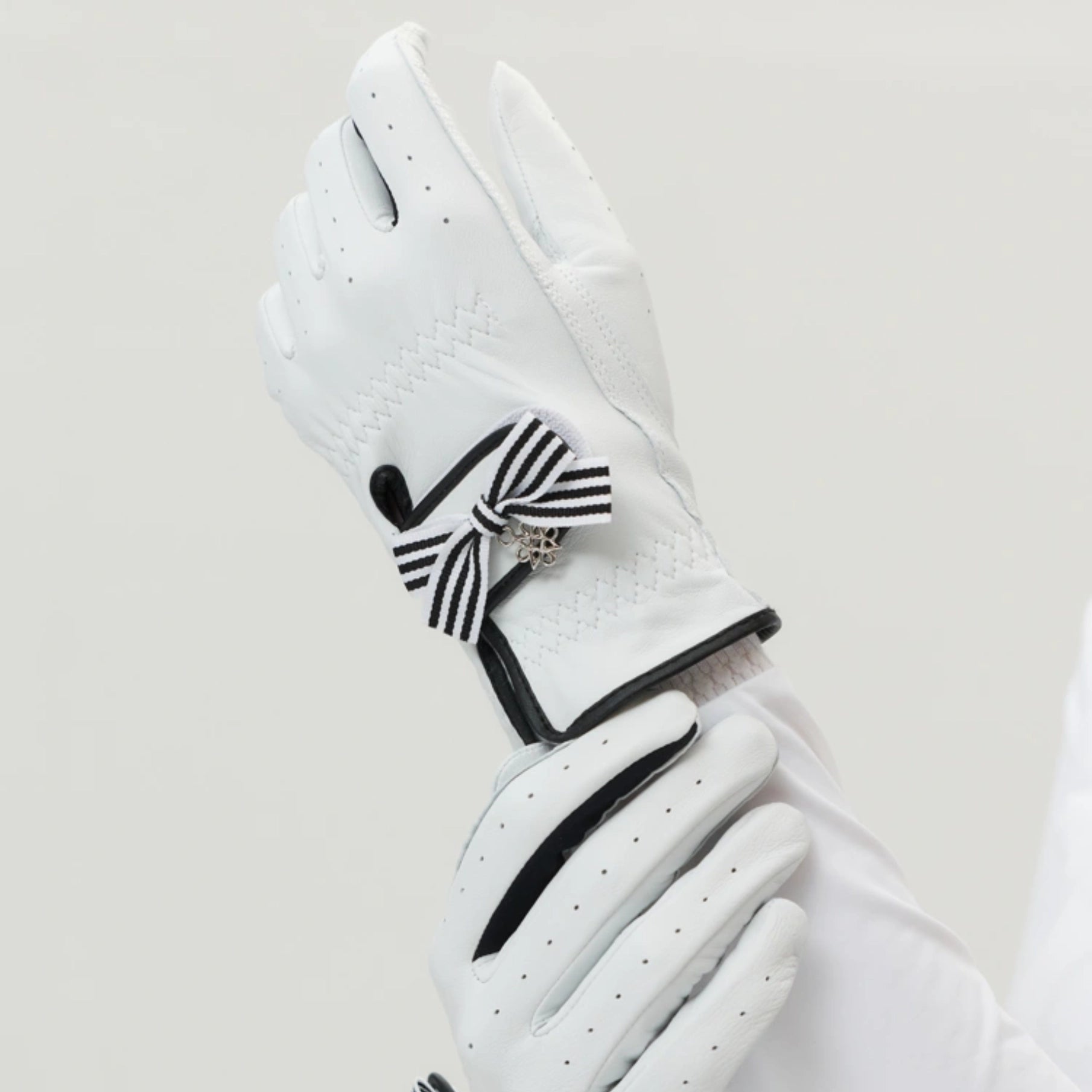 A pair of white leather golf gloves with black trim and a black-and-white striped bow detail on the back of one glove, shown against a light gray background.
