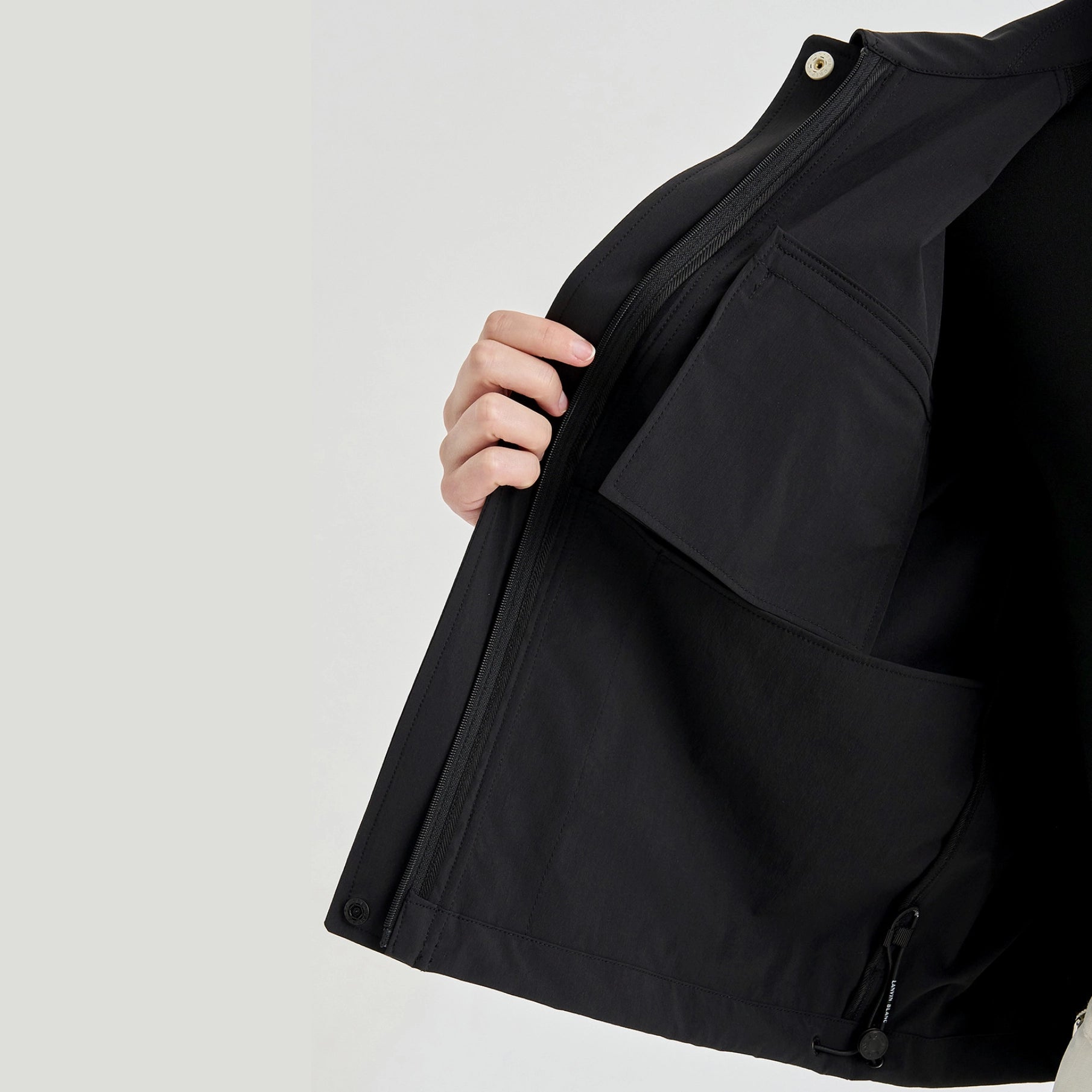 Close-up of a person wearing a black jacket, showing the inner side with a large zip pocket and a hand holding the zipper open, against a light gray background.