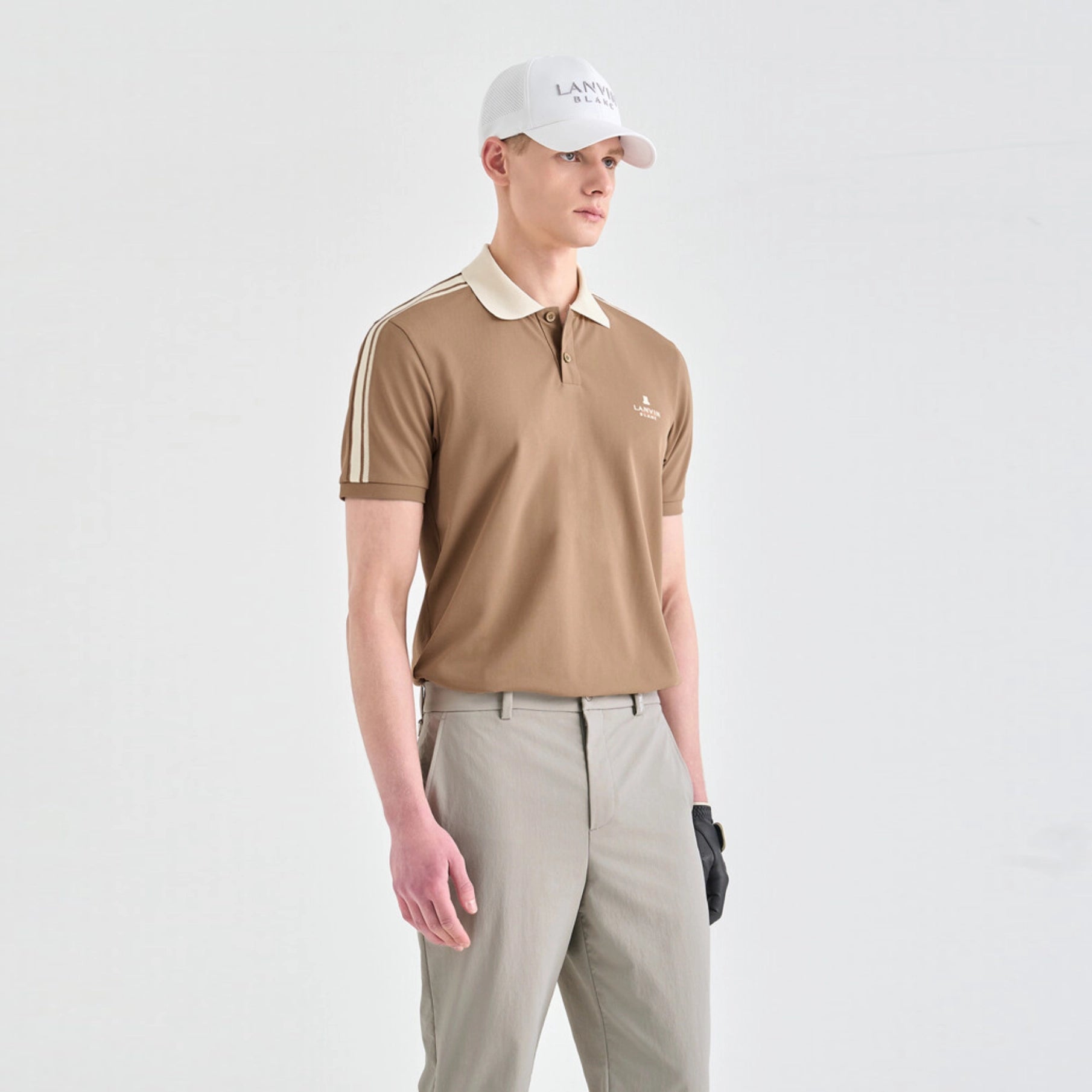 A man modeling a tan short-sleeve polo shirt with a cream collar and three cream stripes on the sleeve, paired with khaki pants and a white cap, against a plain light gray background.