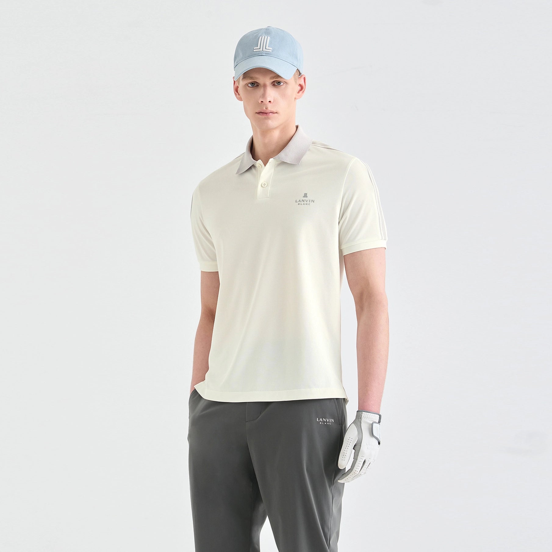 A man modeling a light cream-colored short-sleeve polo shirt with a small embroidered logo on the left chest, paired with dark gray golf pants, a light blue cap, and a white golf glove, against a plain light gray studio background.