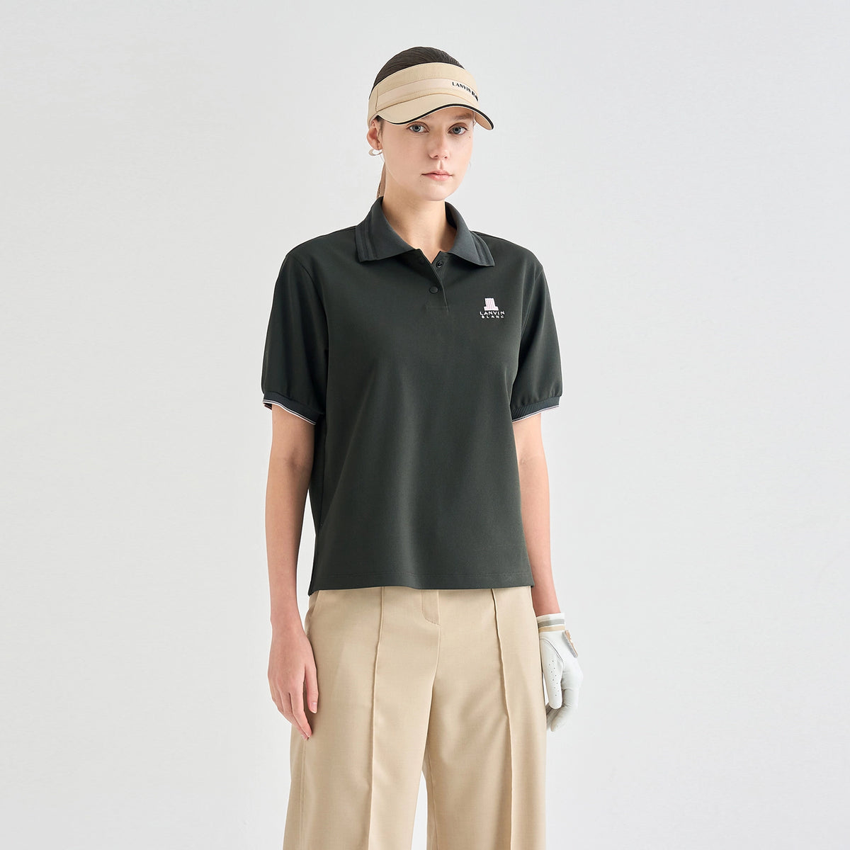 A model wearing a dark olive green short-sleeve polo shirt with a small white embroidered logo on the left chest, paired with beige pants, a beige visor, and a white golf glove, against a light gray studio background.