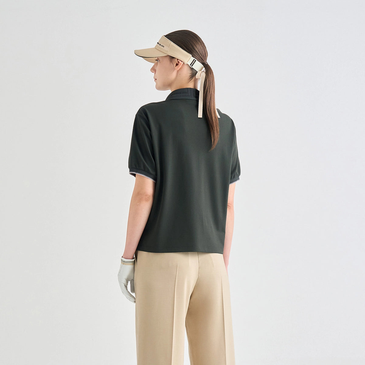 A woman shown from the back wearing a dark olive green short-sleeve polo shirt, beige pants, a beige visor, and a white golf glove, against a plain light gray studio background.