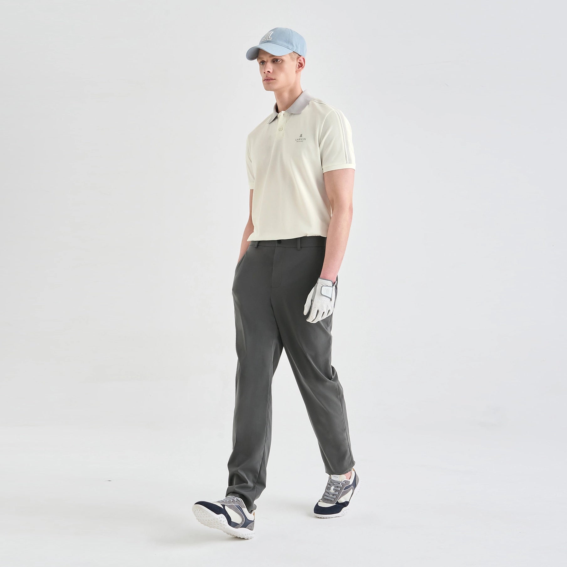 A man modeling a light cream short-sleeve polo shirt, dark gray golf pants, a light blue baseball cap, a white golf glove, and golf shoes against a plain white studio background.