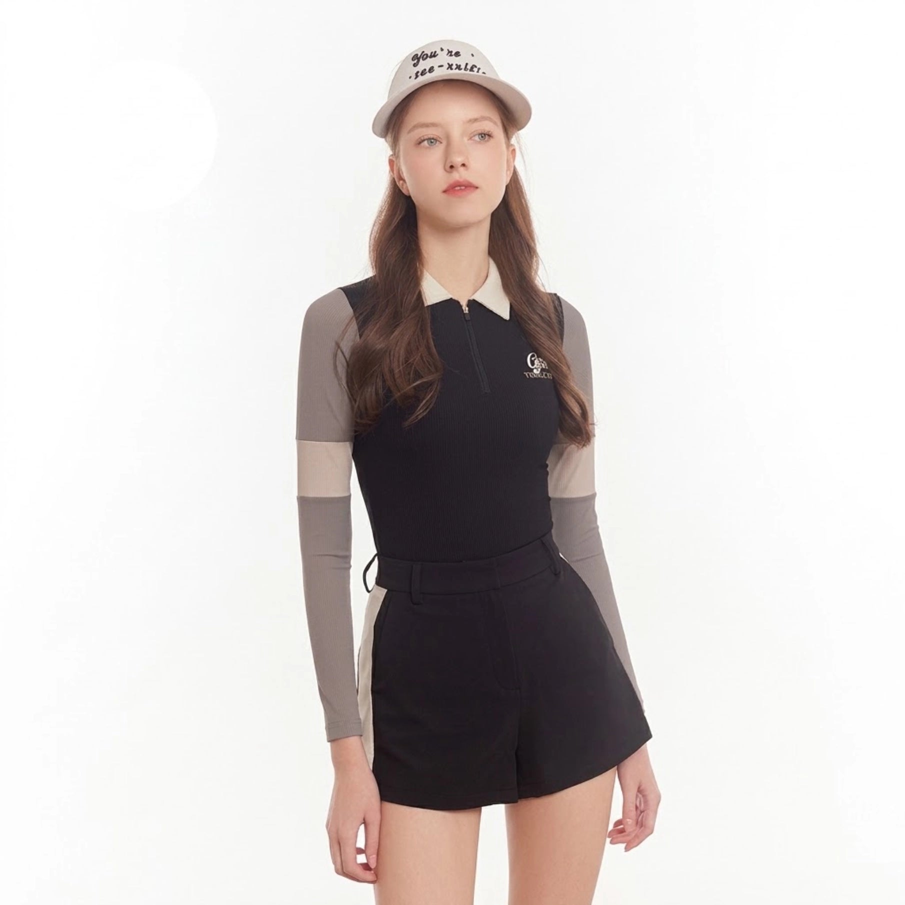 A woman modeling a black long-sleeve collared top with gray sleeves, paired with black shorts and a beige baseball cap, against a plain white background.
