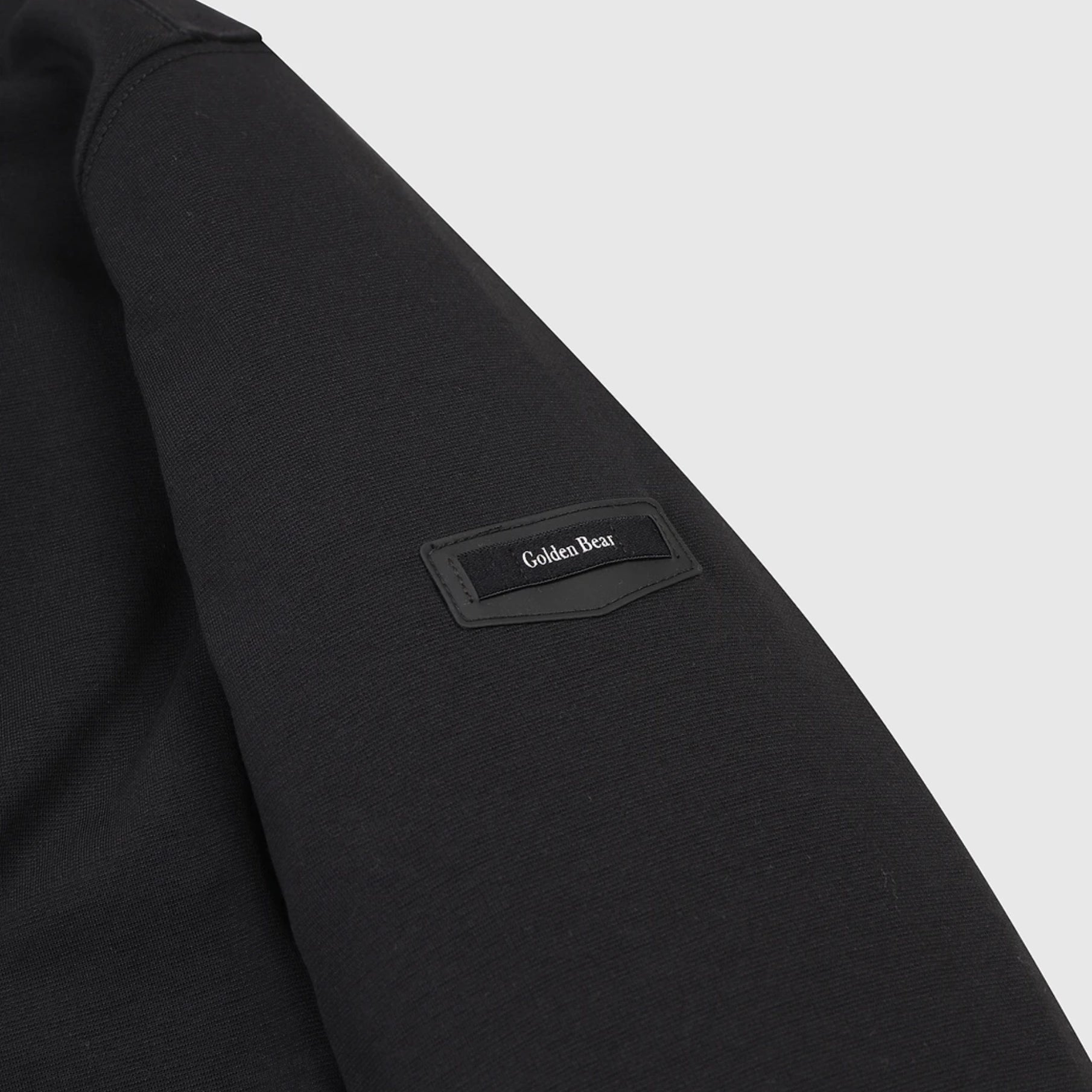 Close-up of a black sweatshirt sleeve with a tonal black patch on the upper arm that reads 'Golden Bear' in white text, against a light gray background.