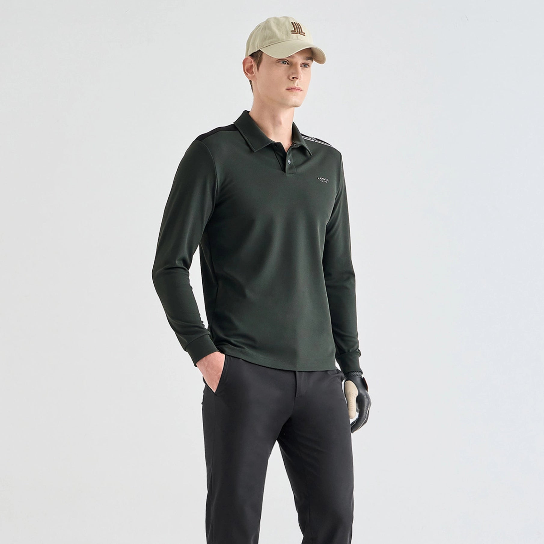 A man modeling a dark green long-sleeve polo shirt, black golf pants, a beige baseball cap, and a black golf glove against a light gray studio background.