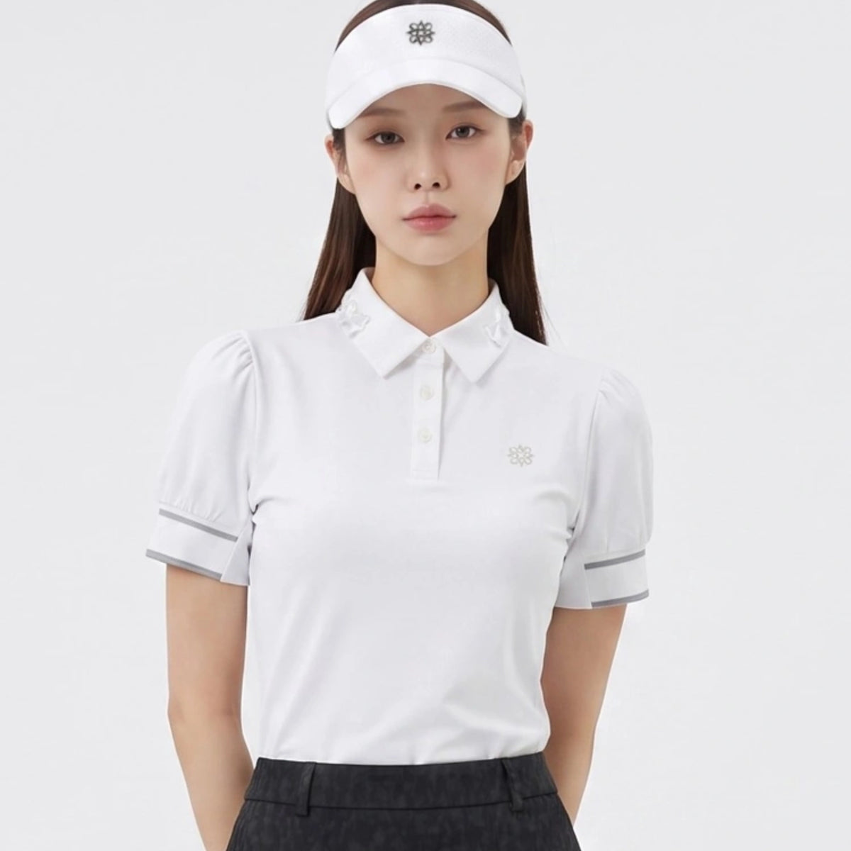 A woman modeling a white short-sleeve polo golf shirt with puffed sleeves, a small embroidered logo on the chest, and a matching white visor, set against a plain light gray studio background.