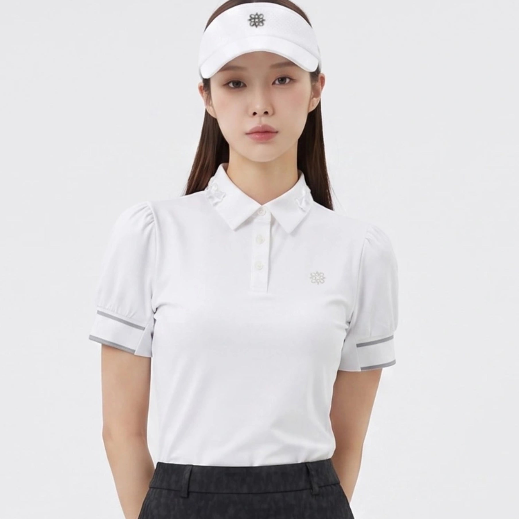 A woman modeling a white short-sleeve polo golf shirt with puffed sleeves, a small embroidered logo on the chest, and a matching white visor, set against a plain light gray studio background.
