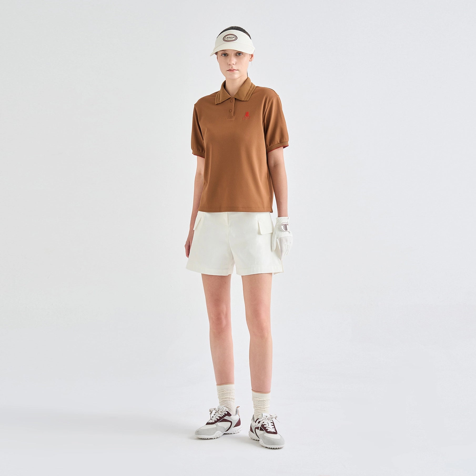 A model stands against a light gray studio background wearing a brown short-sleeve polo shirt with a small red embroidered logo on the chest, off-white shorts, a matching visor, a white glove, white socks, and brown-and-white sneakers.