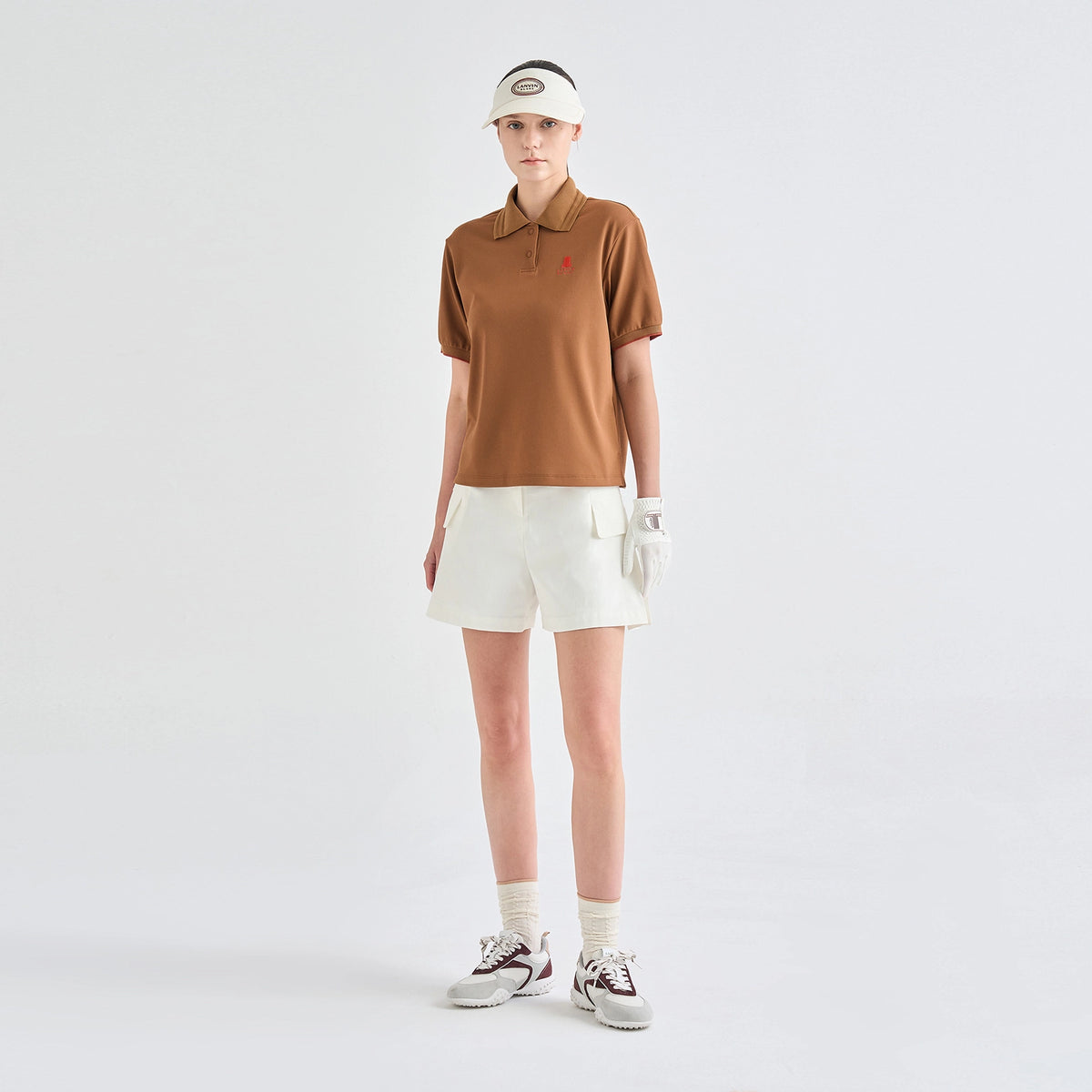 A model stands against a light gray studio background wearing a brown short-sleeve polo shirt with a small red embroidered logo on the chest, off-white shorts, a matching visor, a white glove, white socks, and brown-and-white sneakers.