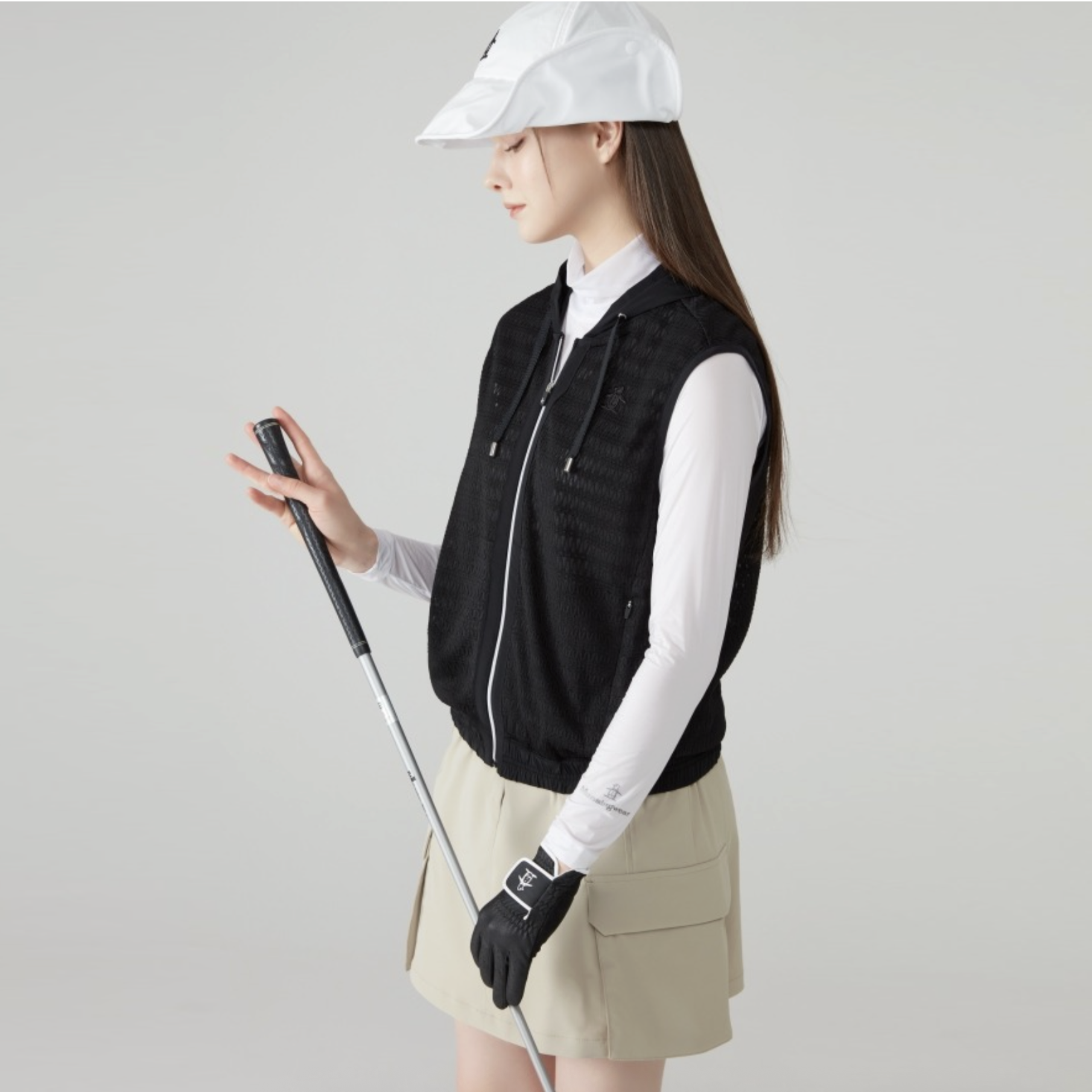 A woman modeling a black zip-up sleeveless vest over a white long-sleeve shirt, paired with a beige mini skirt, black golf glove, and white cap, holding a golf club against a light gray studio background.