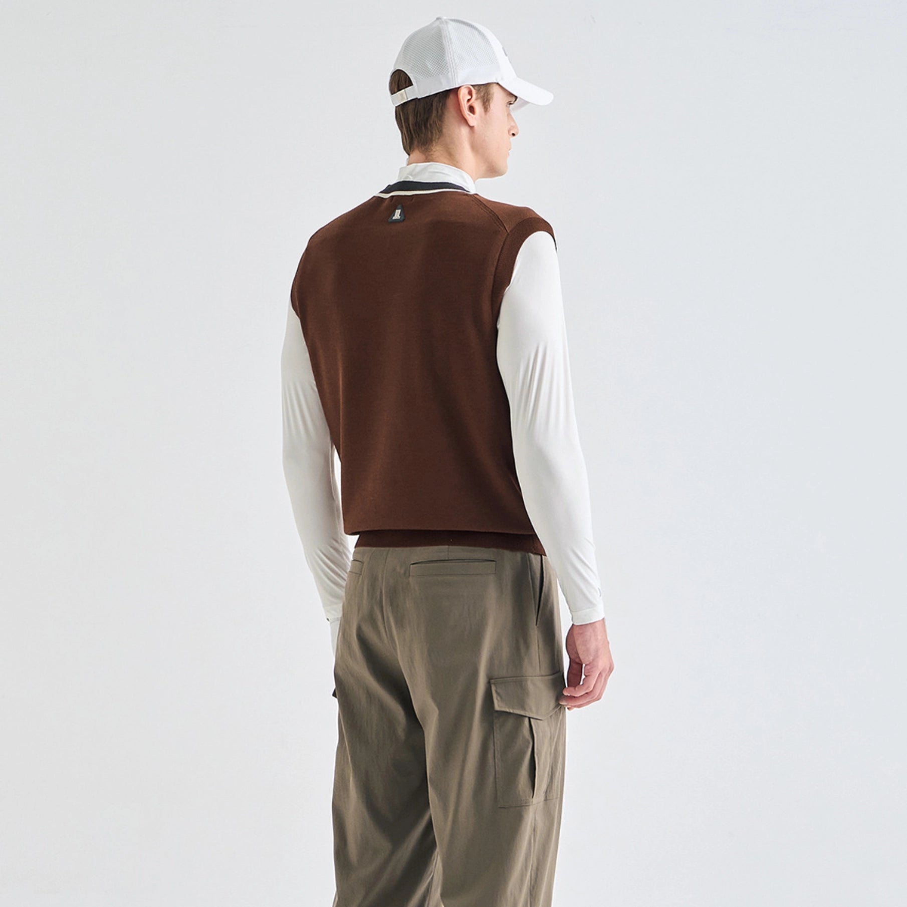 A man shown from the back wearing a brown knit sweater vest over a white long-sleeve shirt, paired with olive cargo pants and a white baseball cap, against a plain light gray studio background.