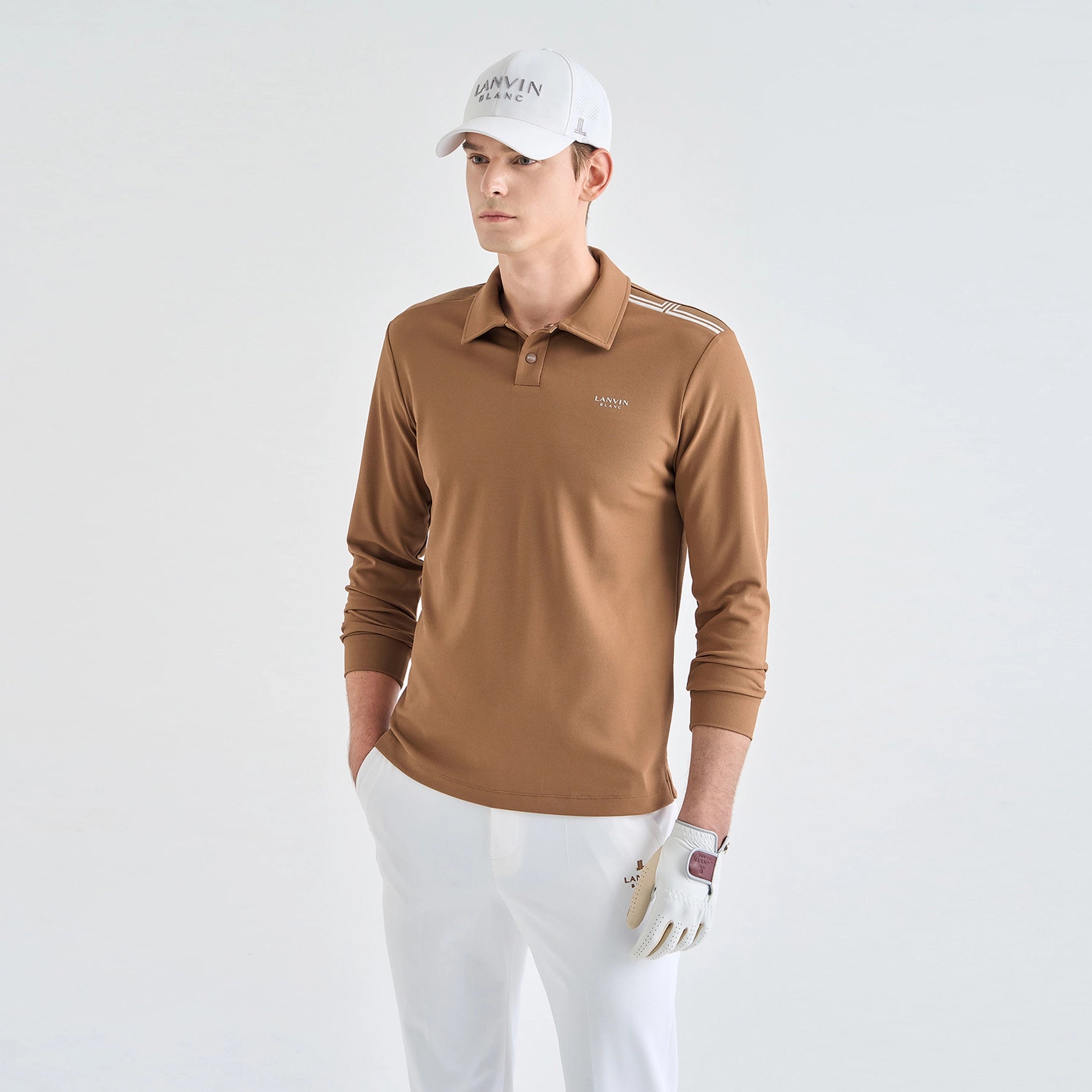 A man modeling a long-sleeve brown polo shirt with white golf pants, a white cap, and a white golf glove against a plain light gray studio background.