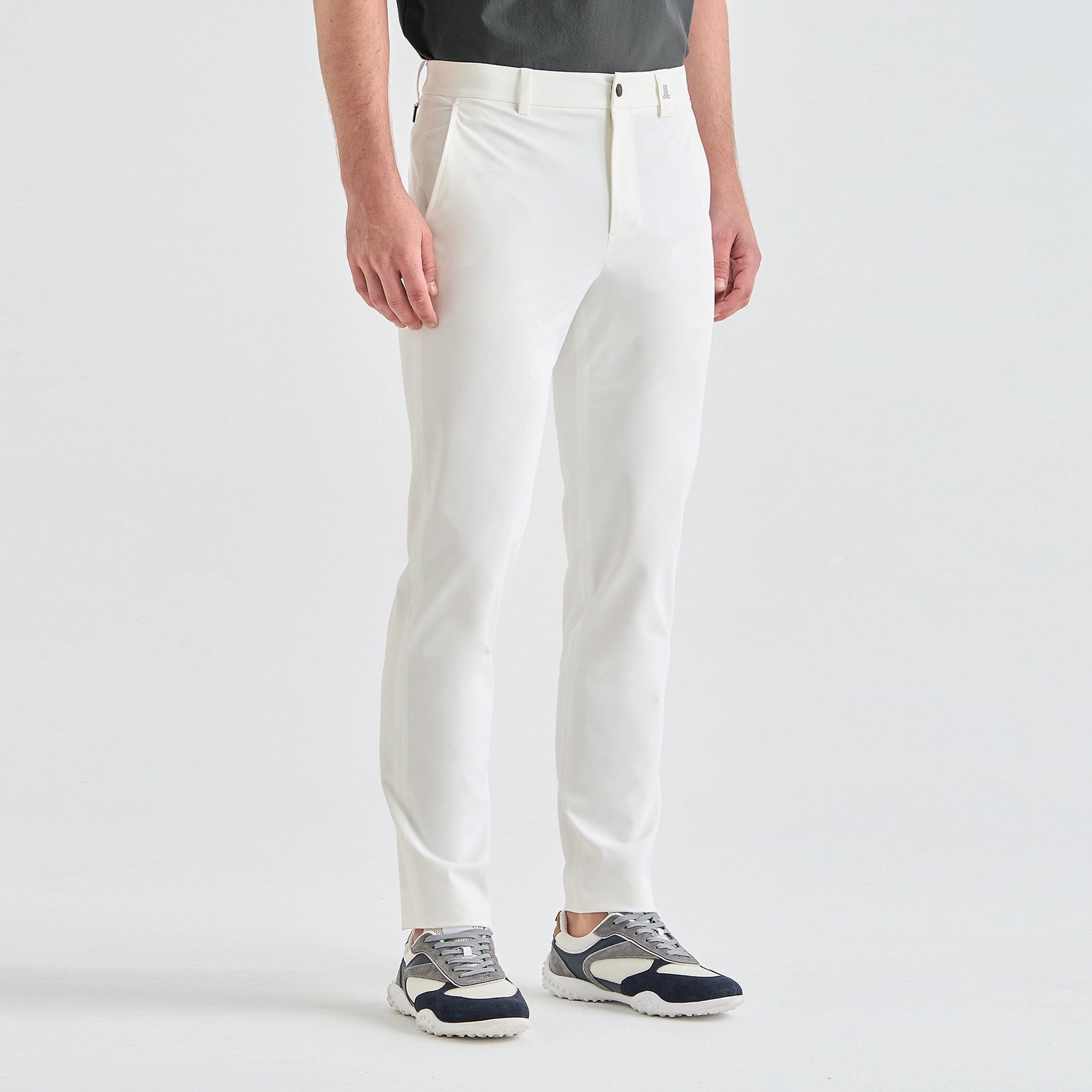 A model from the waist down wearing slim-fit white trousers and dark gray and navy sneakers, standing against a clean white studio background.