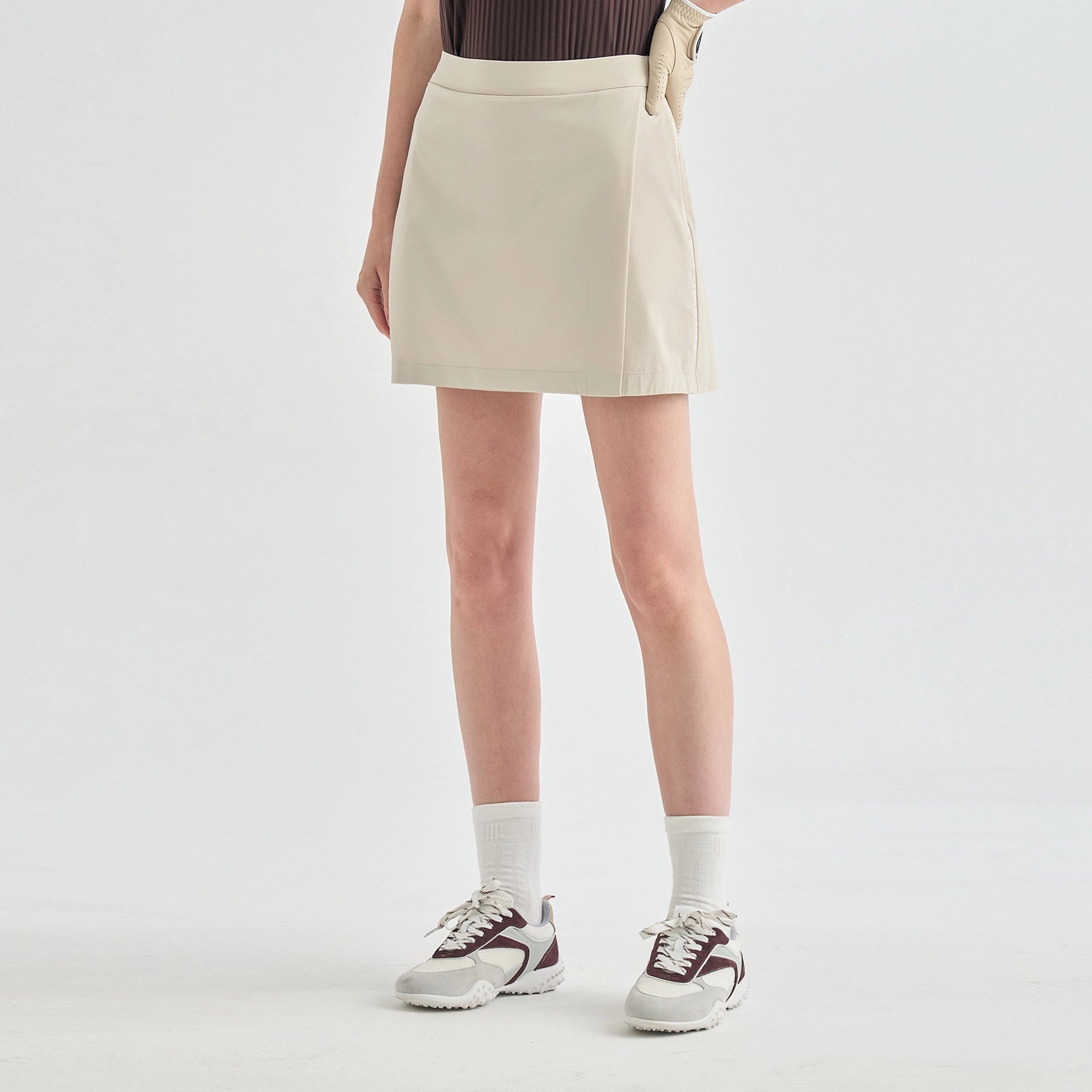 A model wearing a light beige athletic mini skirt, paired with white crew socks and white sneakers with dark accents, shown from the waist down against a light gray studio background.