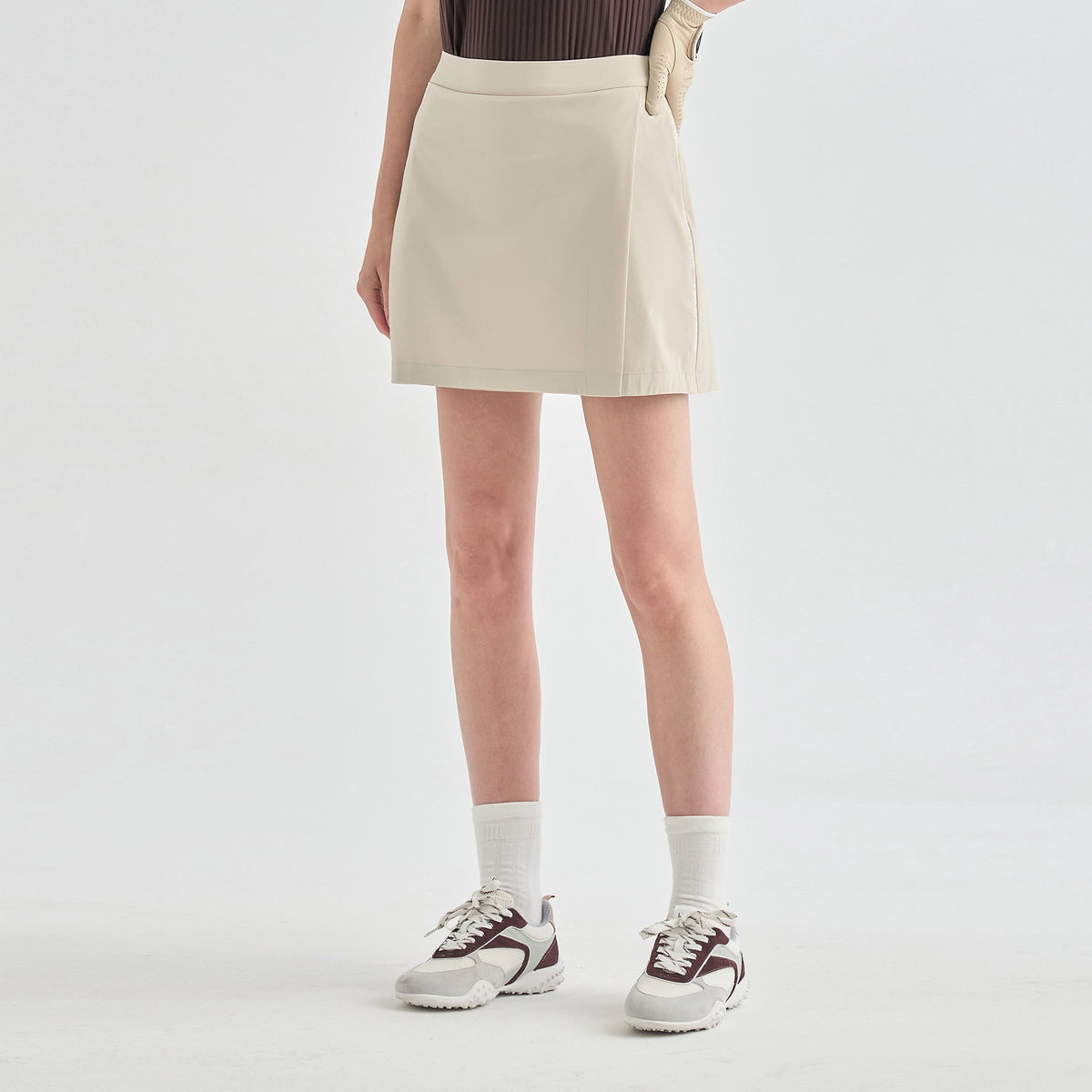 A model wearing a light beige athletic mini skirt, paired with white crew socks and white sneakers with dark accents, shown from the waist down against a light gray studio background.