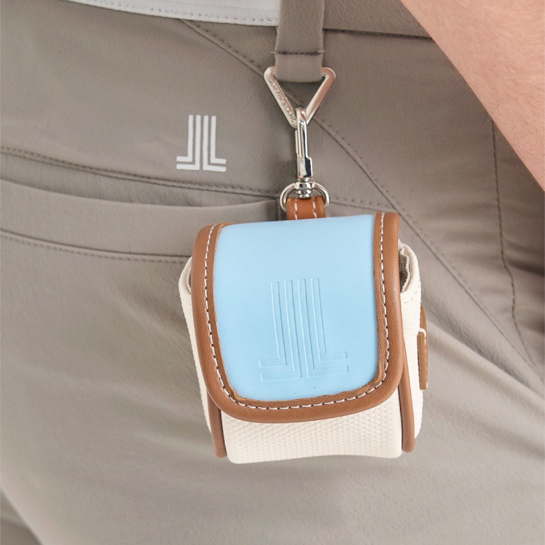 A small light blue and off-white pouch with tan trim hanging from a silver carabiner on a person's khaki pants.