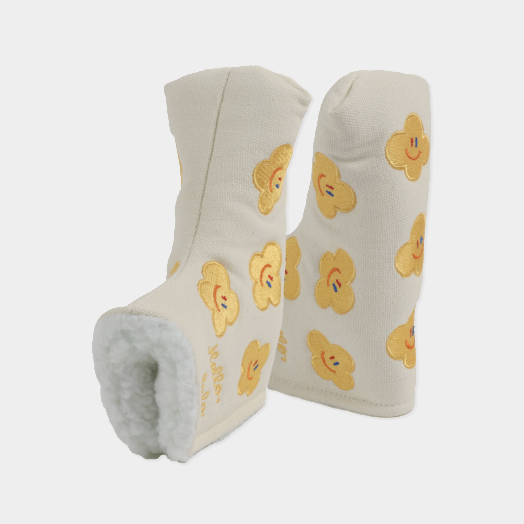 A pair of cream-colored headcovers with a soft white fleece lining and yellow embroidered smiling flower patterns, shown against a light gray background.